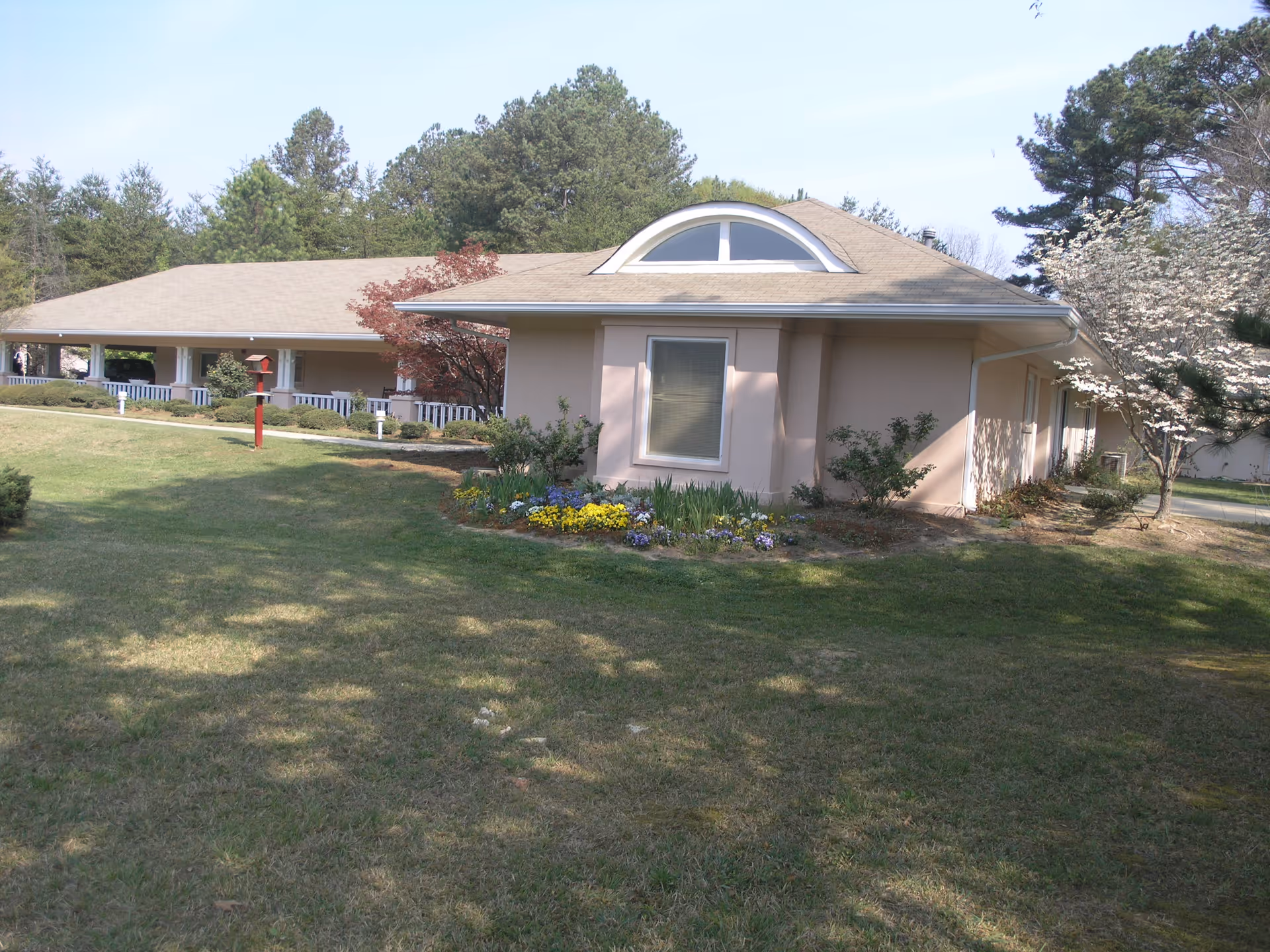 Exterior of a single-story building with a manicured lawn, flowerbeds, and surrounding trees under a clear sky.