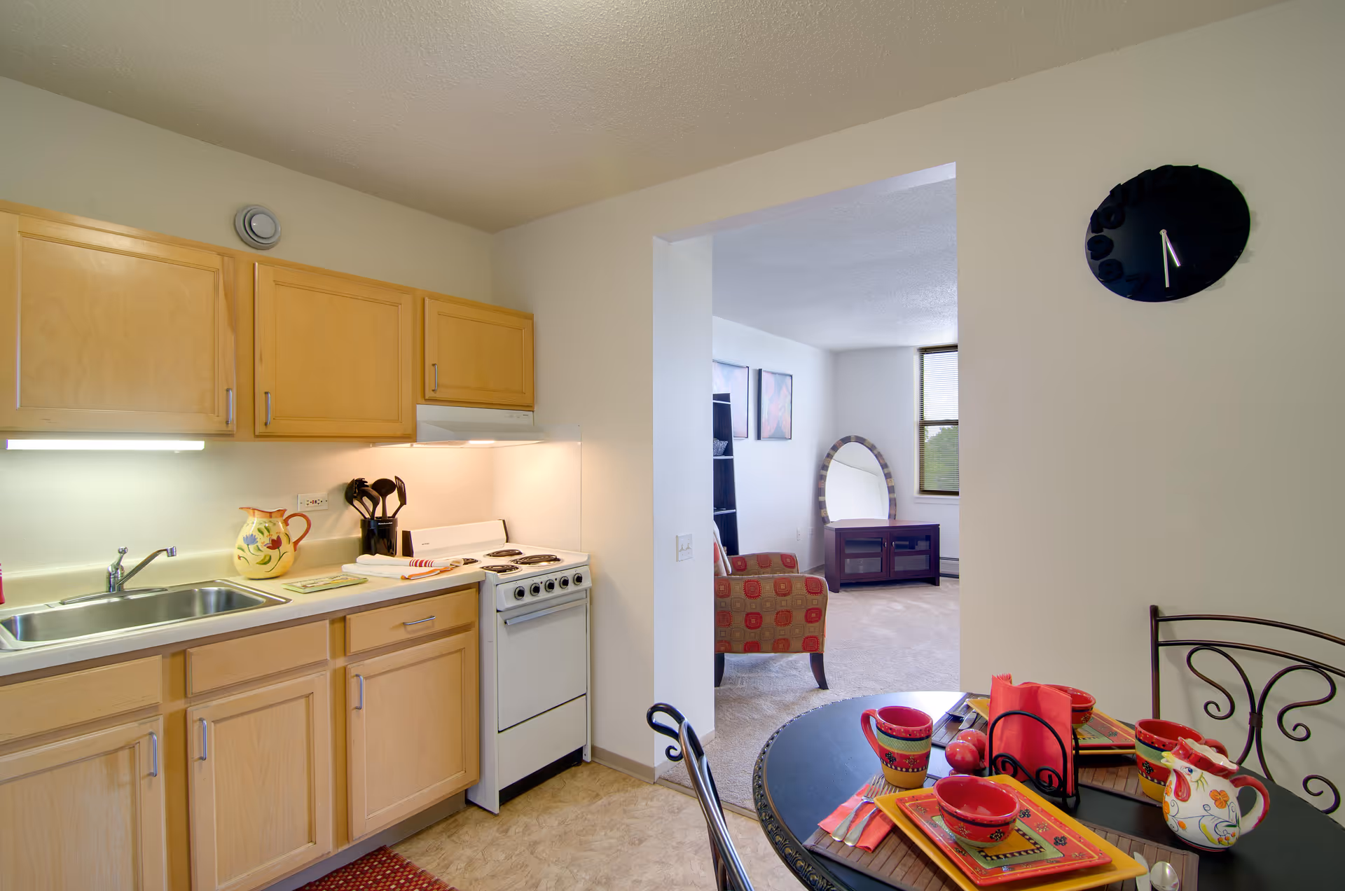 A small kitchen area with light wood cabinets, a stainless steel sink, a white stove, and a countertop with kitchen utensils and a decorative pitcher. Adjacent to the kitchen is a dining table set with colorful plates, bowls, cups, and napkins. Beyond the kitchen and dining area, a living room is visible with a red patterned armchair, a round mirror on a stand, a TV stand, and two framed pictures on the wall.