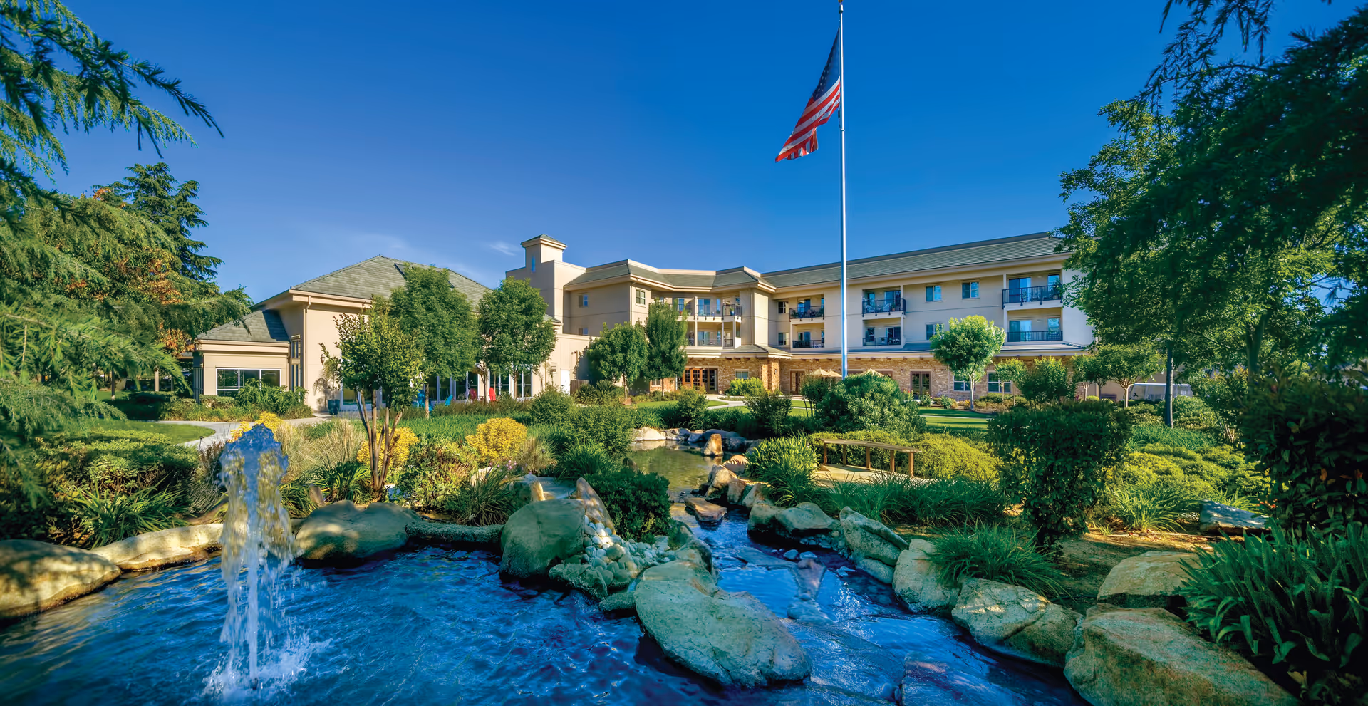 Exterior view of Covenant Living of Turlock facility with a landscaped garden featuring a pond, rocks, and a water fountain in the foreground. The building is a multi-story structure with balconies, surrounded by trees and greenery under a clear blue sky. An American flag is prominently displayed on a flagpole near the center of the image.