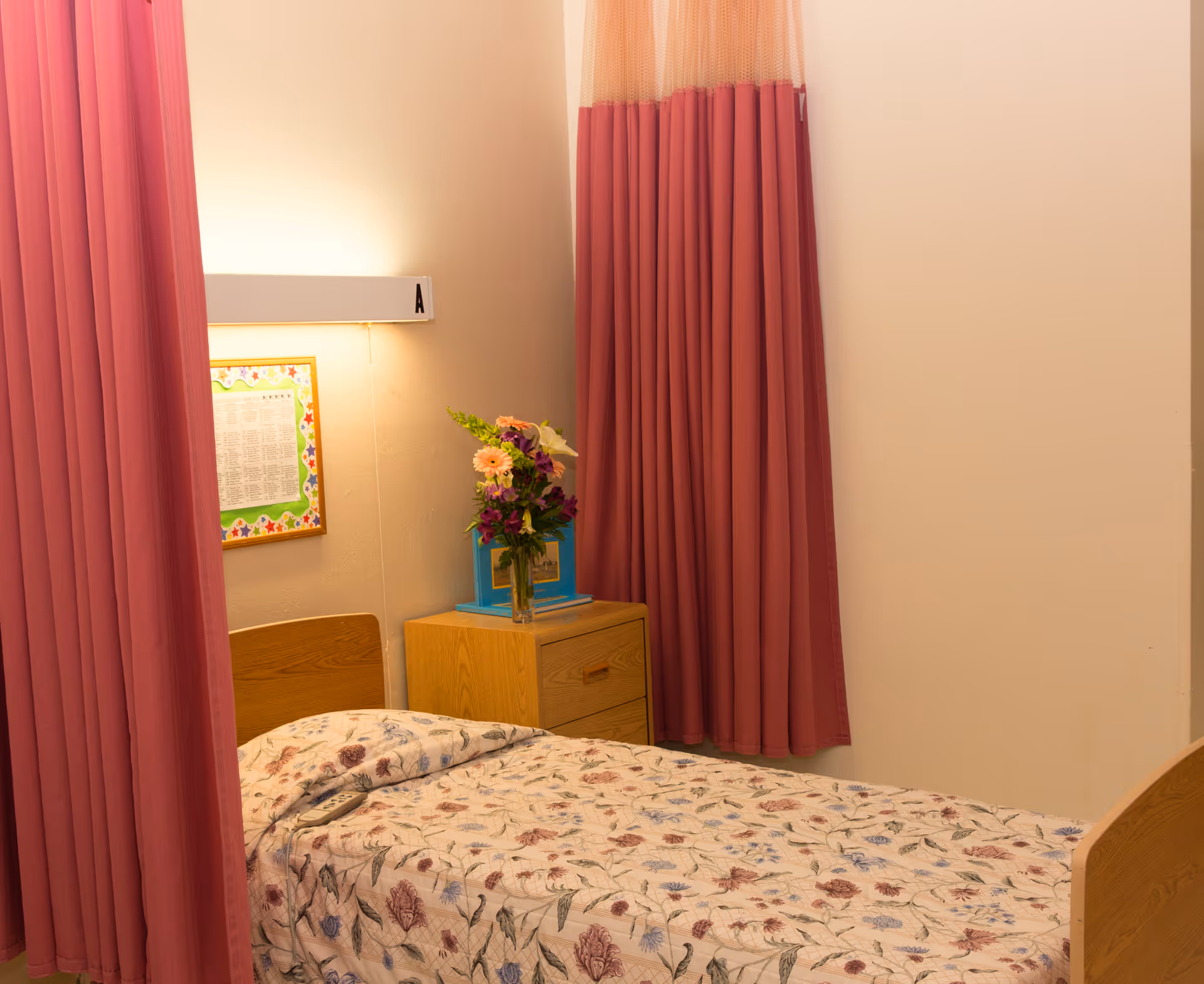 A single bed with a floral bedspread next to a wooden nightstand with a vase of flowers, framed wall art, and pink privacy curtains.