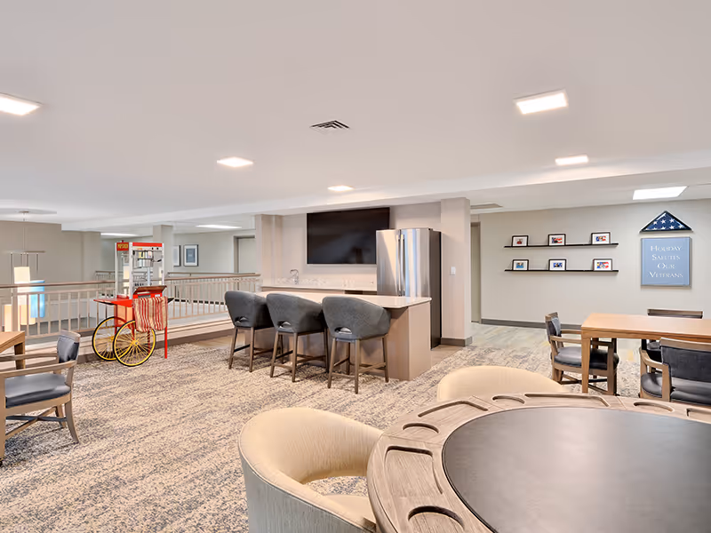 A spacious senior living community common area featuring a kitchen island with four bar stools, a stainless steel refrigerator, a wall-mounted TV, several tables and chairs, a popcorn machine on wheels, and a wall display with framed photos and a sign that reads 'Holiday Salutes Our Veterans.'