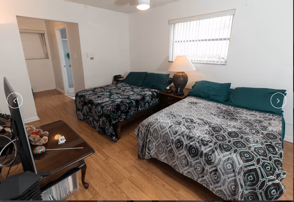 A bedroom in Margate Assisted Living featuring two double beds with patterned bedspreads and green pillows. Between the beds is a wooden nightstand with a lamp and a phone. A window with vertical blinds is above the nightstand. A TV is positioned on a wooden table opposite the beds, with a teddy bear and some small items on the table. The room has wooden flooring and white walls, with a doorway leading to another room or bathroom.