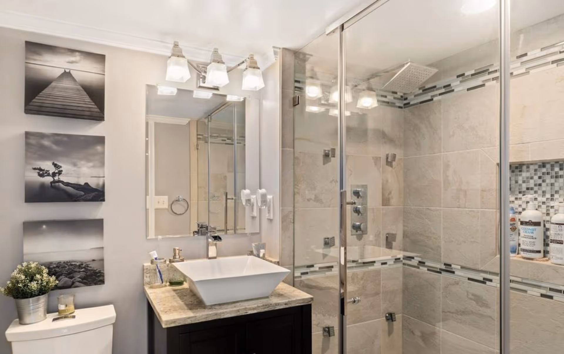 Modern bathroom with a glass-enclosed shower featuring beige tiles and a mosaic tile accent strip. A white vessel sink sits on a granite countertop with a dark wood vanity. Above the sink is a large mirror with three light fixtures. On the left wall, there are three black and white framed photos and a small plant on top of the toilet tank.