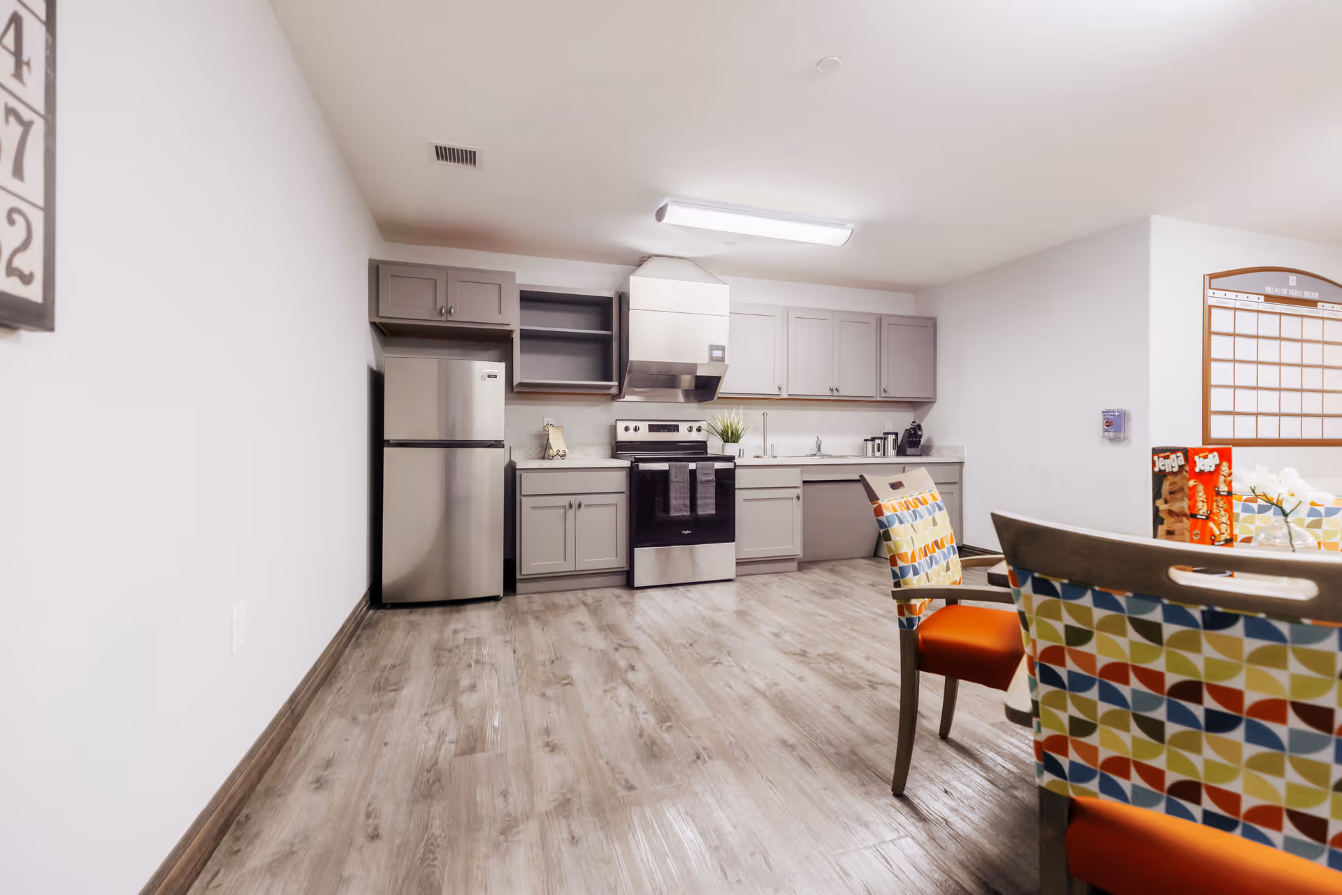 A modern kitchen area with light gray cabinets, a stainless steel refrigerator, stove, and range hood. The floor is wood-style laminate, and there are colorful patterned chairs with orange seats around a table on the right side. A large wall calendar and some board games are visible on the far right wall.