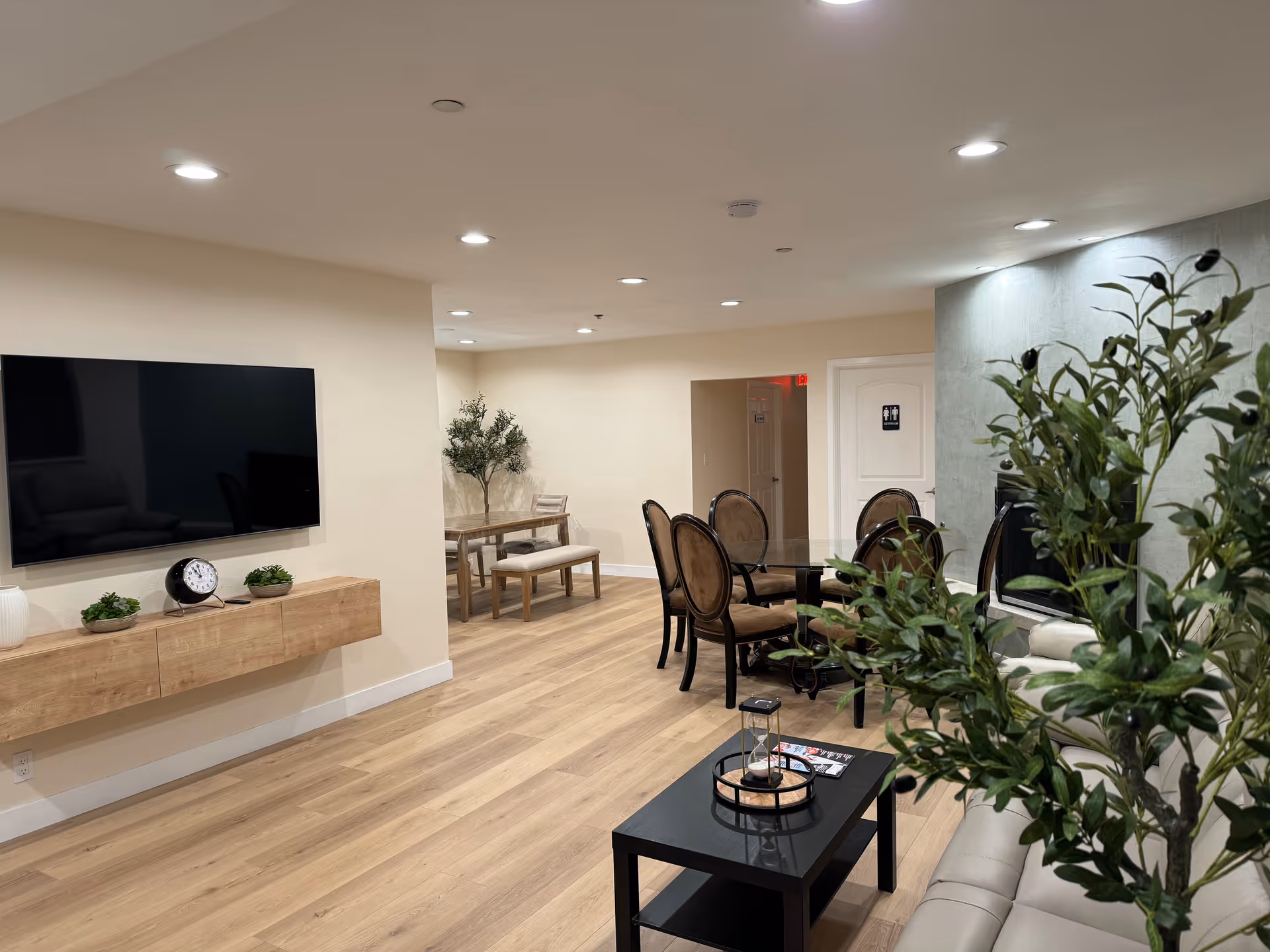 A spacious senior living common area with light wood flooring and recessed ceiling lights. The room features a wall-mounted flat-screen TV above a floating wooden shelf with decorative items, a glass-top dining table with six upholstered chairs, a smaller wooden table with a bench and chair in the background, and a beige leather sofa partially visible behind a large green plant in the foreground. A door with a restroom sign is visible in the back.