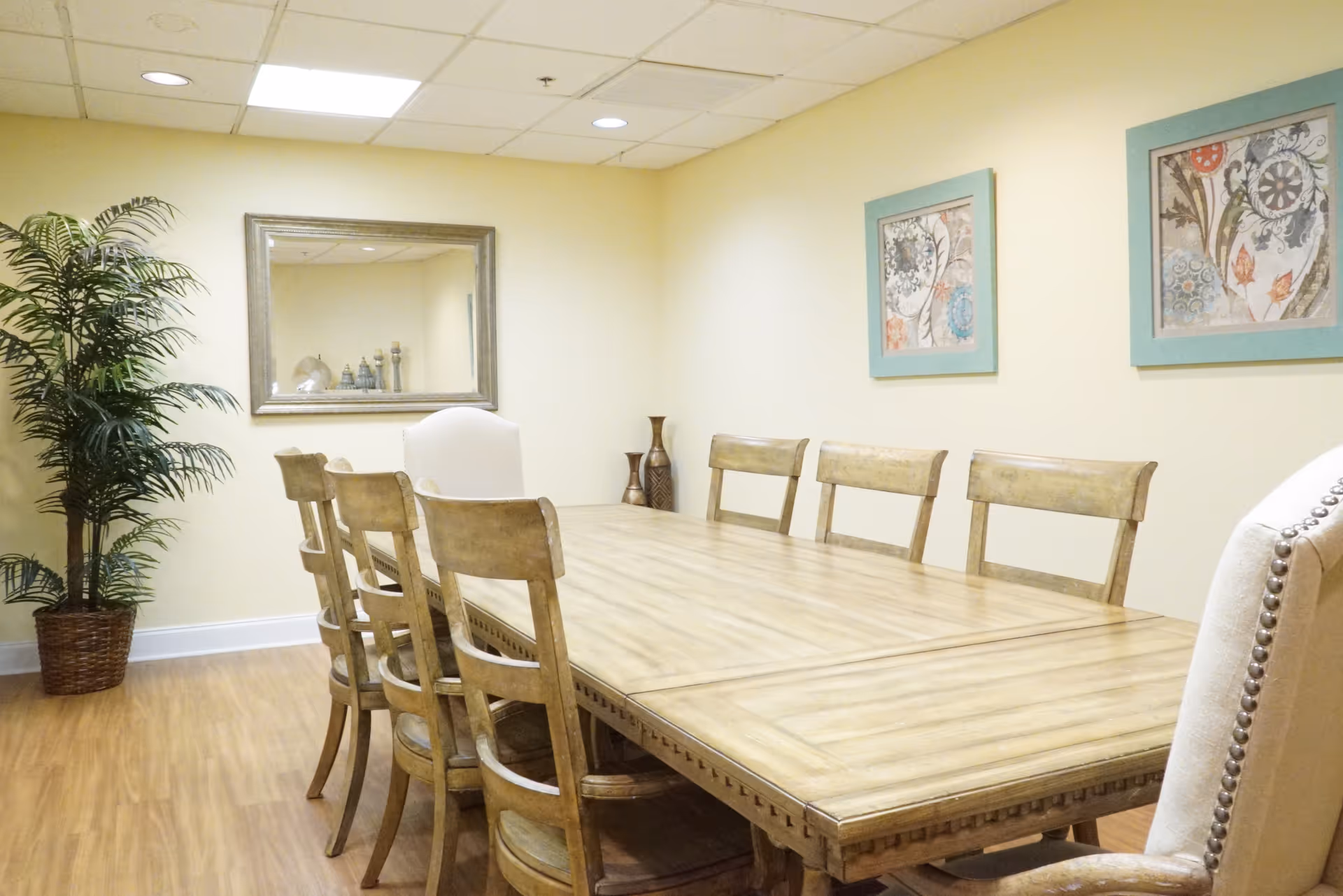 A dining room with a long wooden table surrounded by eight chairs, two of which have white upholstery with nailhead trim. The room has light yellow walls, a large mirror on one wall, two framed floral artworks on another wall, a potted plant in the corner, and wooden flooring.