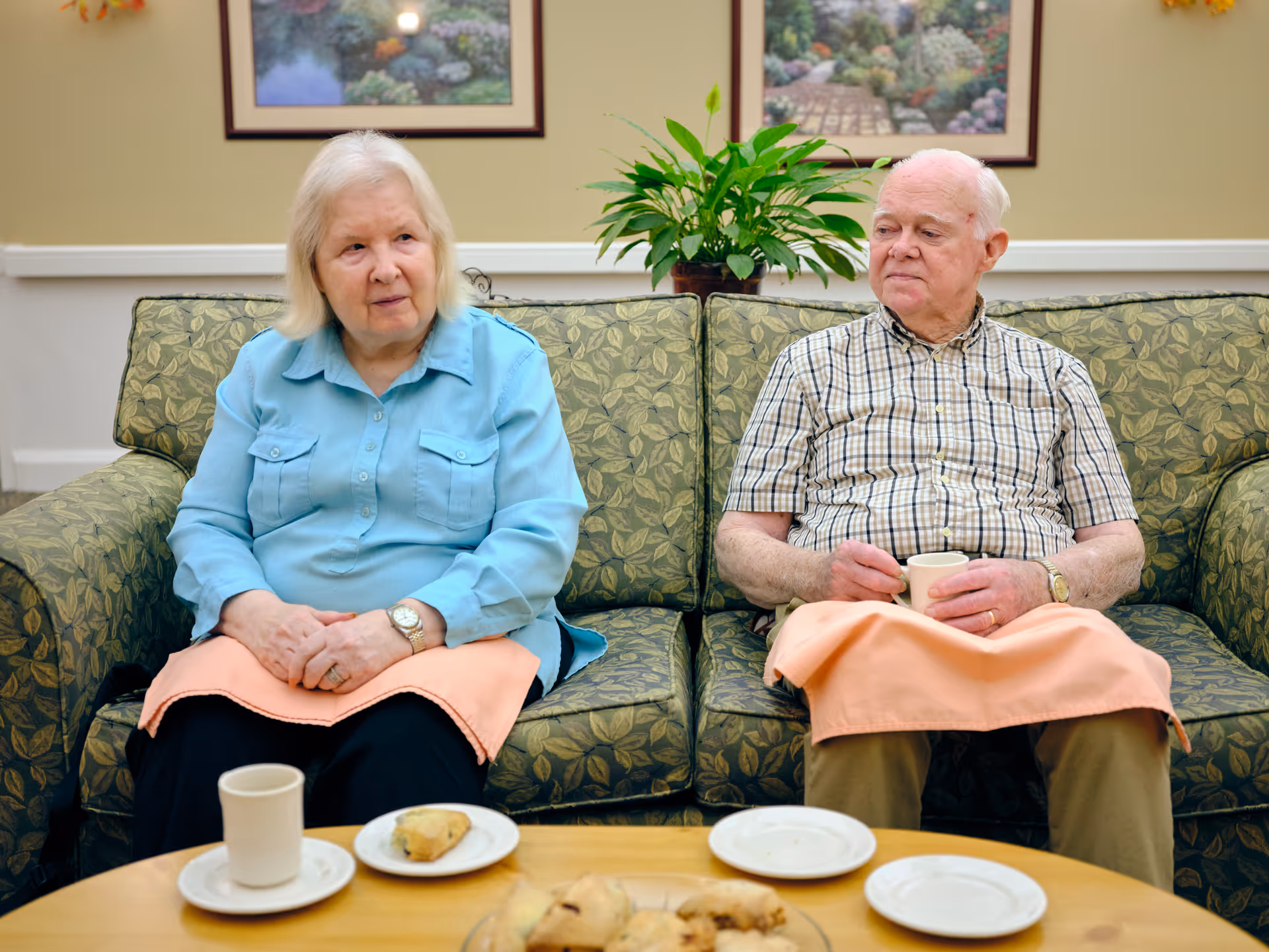 An elderly woman and man sitting on a green patterned couch in a living room setting. Both have peach-colored napkins on their laps. The woman is wearing a light blue shirt and the man is wearing a plaid shirt. They each have a cup in front of them on a wooden table with plates and pastries. There are two framed pictures and a green plant on a shelf behind them.