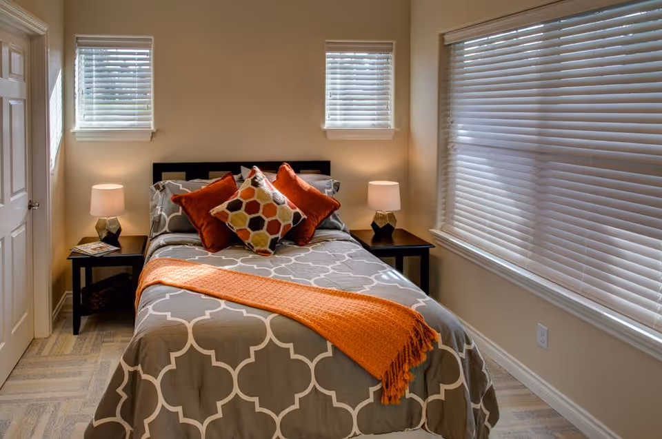 A cozy bedroom with a bed centered between two small windows. The bed has a gray and white patterned comforter, three decorative pillows in orange and multicolor hexagon patterns, and an orange throw blanket. There are two black nightstands on either side of the bed, each with a lamp. A large window with white blinds is on the right wall, and a closed door is on the left.