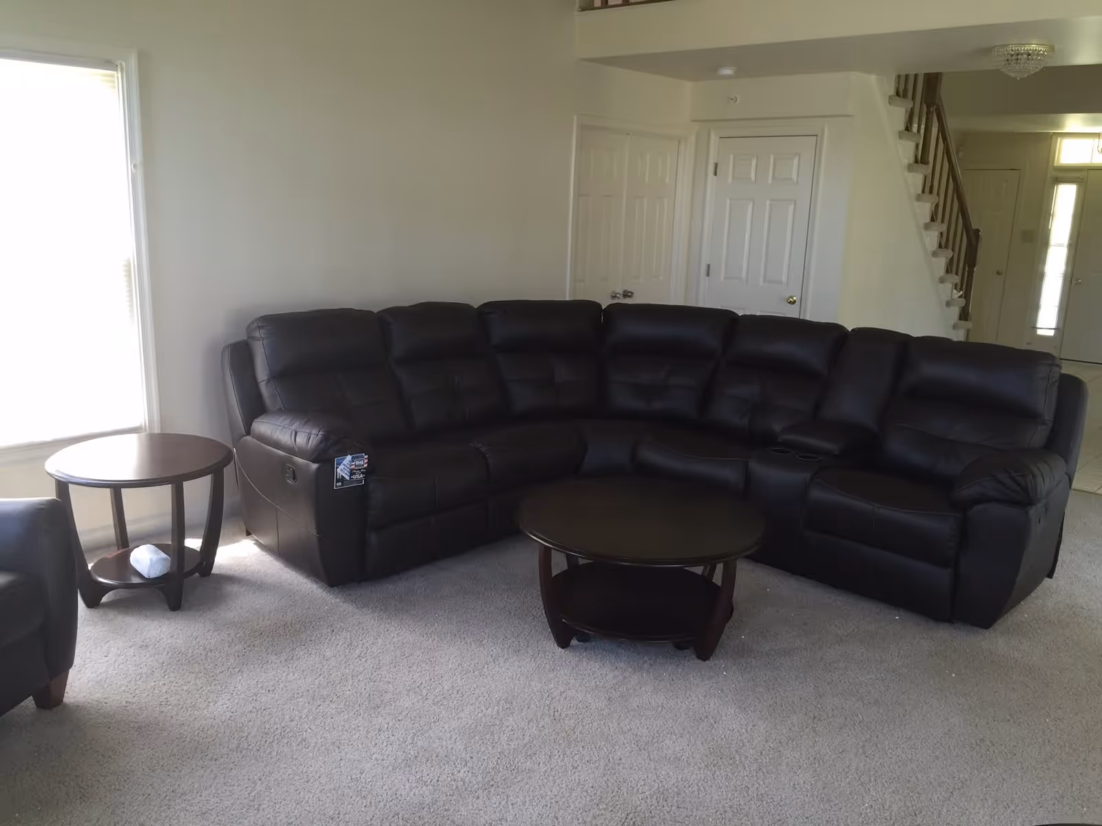 A spacious living room with a large black leather sectional sofa, a round wooden coffee table in front of it, and a matching round side table next to the sofa. The room has light-colored carpet and walls, with a window letting in natural light on the left. In the background, there are two white doors and a staircase leading to an upper floor.