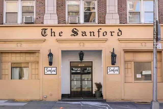 Exterior front entrance of a building named The Sanford with beige walls, two black lantern-style lights flanking the doorway, and two signs reading 'Sanford' on either side of the entrance.