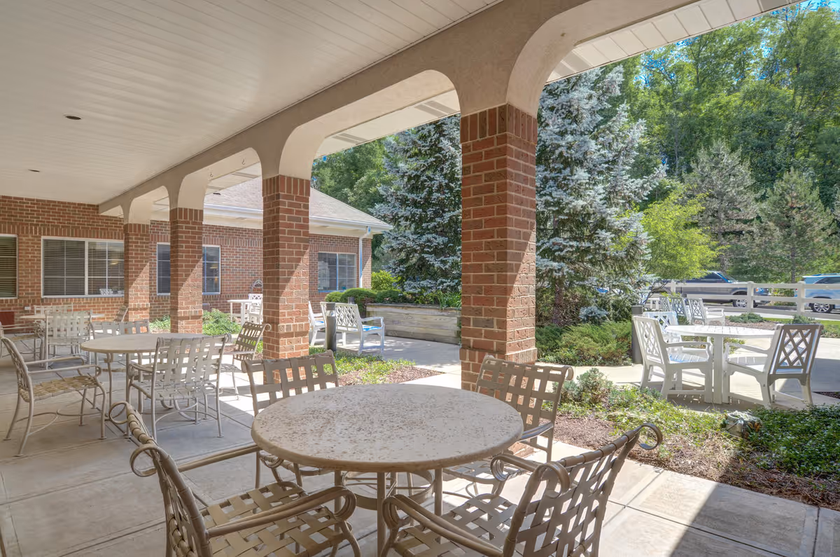 Outdoor covered patio area with multiple round metal tables and chairs. The patio has brick columns and overlooks a garden with trees and shrubs. There are additional tables and chairs outside the covered area, with a parking lot and cars visible in the background.
