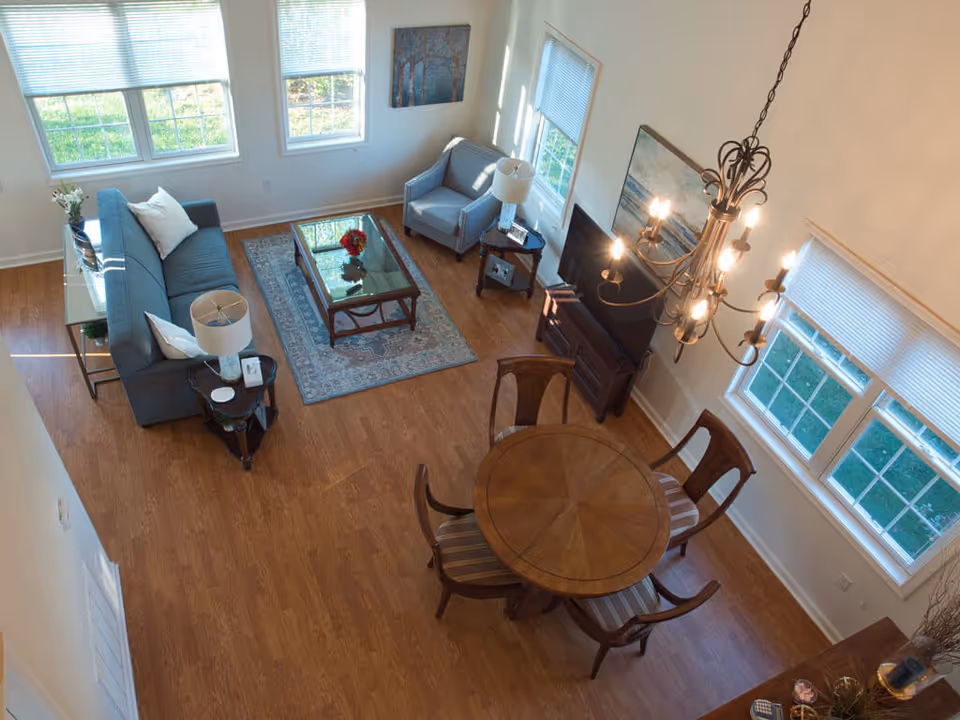 A bright living room with wooden flooring, featuring a blue sofa with white pillows, a glass coffee table on a patterned rug, a blue armchair, a side table with a lamp, a flat-screen TV on a wooden stand, a round wooden dining table with four chairs, and large windows letting in natural light.