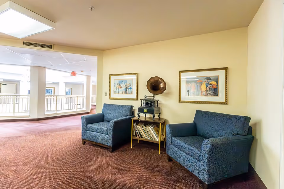 A cozy seating area in a senior living facility hallway with two blue patterned armchairs and a small wooden table between them holding a vintage gramophone and a collection of vinyl records. The walls are light yellow, adorned with two framed paintings, and the floor is covered with a reddish-brown carpet. The hallway is bright with natural light coming from the left side.