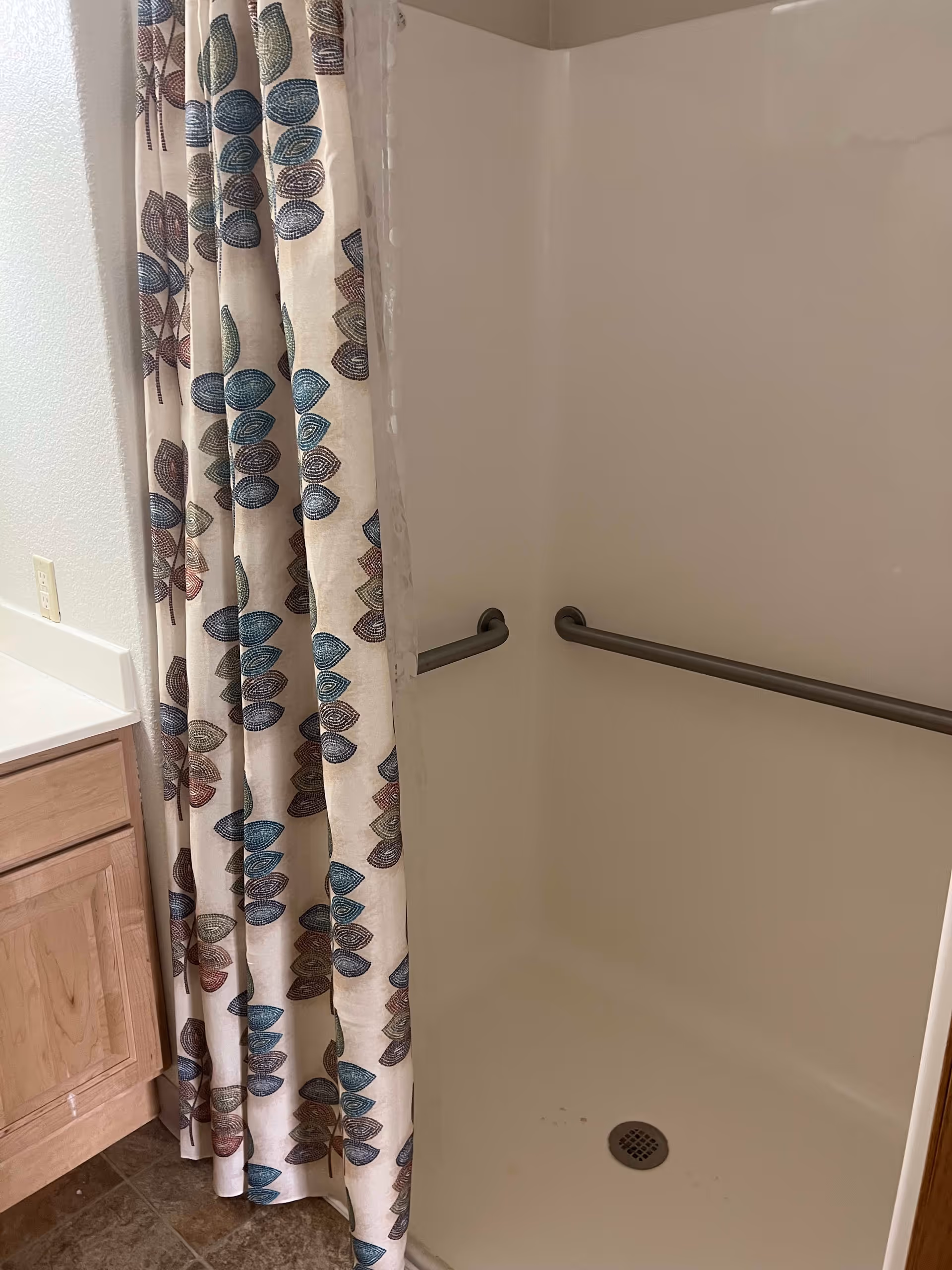 A shower area with a white interior and two horizontal metal grab bars on the walls. A shower curtain with a leaf pattern in blue, green, and brown colors is partially drawn to the side. Next to the shower is a wooden cabinet with a white countertop and a wall outlet above it.