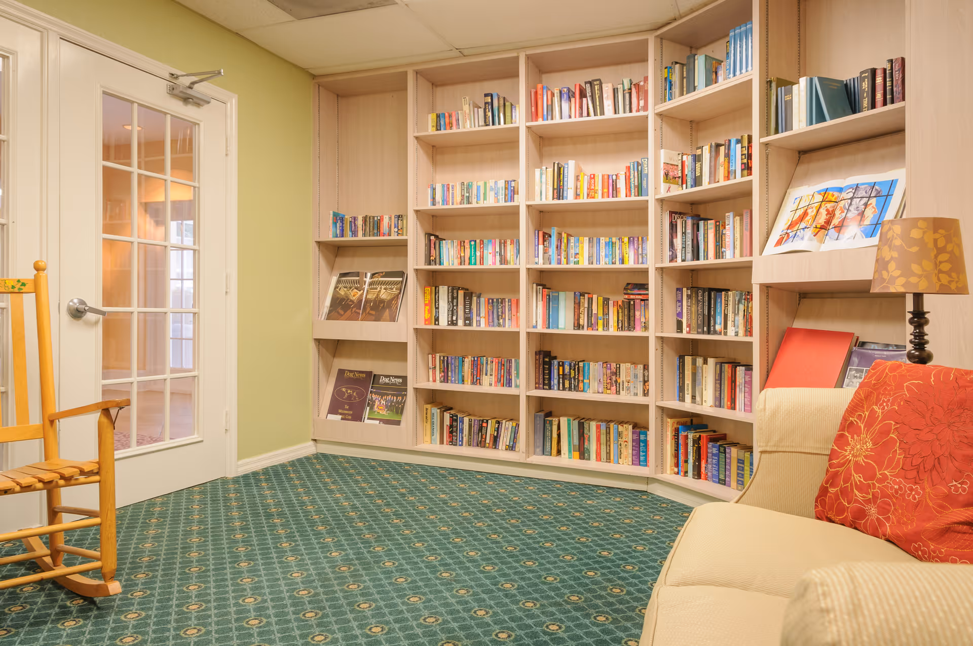 Cozy reading room with built-in wooden bookshelves filled with books, a beige couch with red floral pillows, a wooden rocking chair, a lamp with a patterned shade, and a door with glass panes leading to another room.