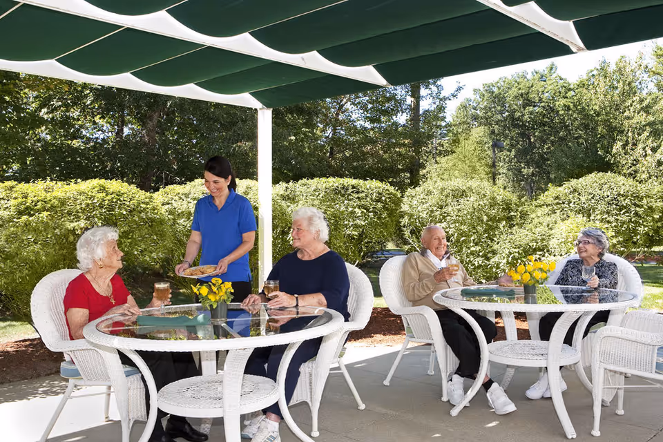 Four elderly people sitting at two white wicker tables with glass tops outdoors under a green canopy. A caregiver in a blue shirt is serving cookies to two elderly women at one table. The elderly people are enjoying drinks and smiling, surrounded by green bushes and trees in a garden setting.