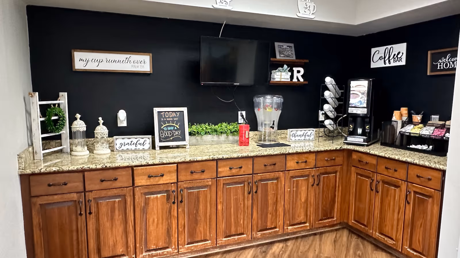 A coffee bar area with wooden cabinets and granite countertops. The back wall is painted black and decorated with various signs including 'Coffee Bar', 'welcome HOME', and inspirational quotes. There is a water dispenser, a coffee machine, and a rack holding coffee pods. Small decorative plants and items are placed on the countertop and shelves.
