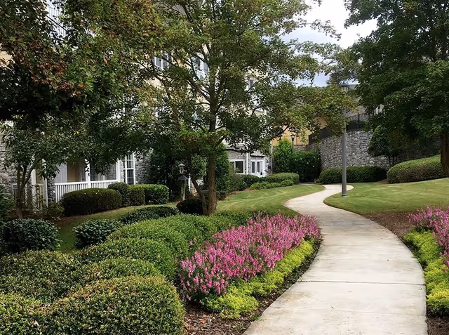 A winding concrete pathway through a landscaped garden area with green bushes, pink flowers, and trees, adjacent to a stone building with white-framed windows and a porch.