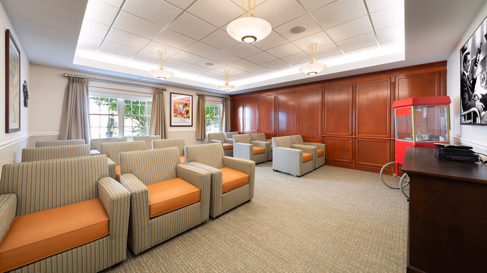 A well-lit lounge area with multiple cushioned armchairs arranged in rows facing a large flat-screen TV mounted on the wall. The room features large windows with beige curtains, wood-paneled walls, and a red popcorn machine on wheels near a wooden cabinet.