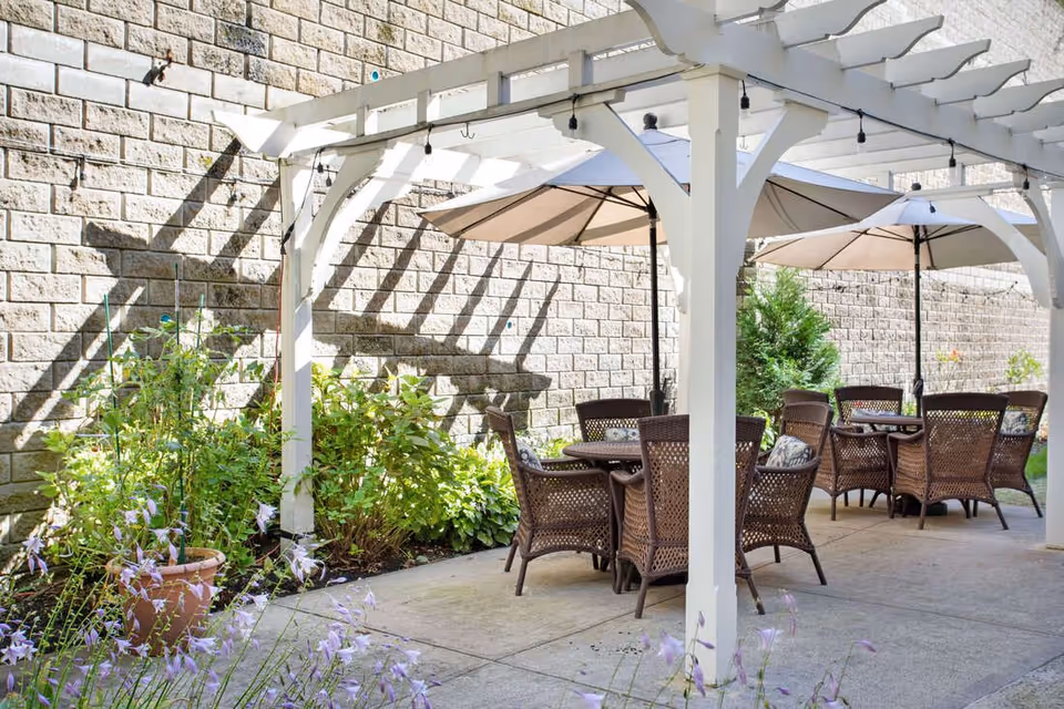 Outdoor patio area with a white pergola providing partial shade over several round tables surrounded by wicker chairs. Large beige umbrellas are open above the tables. There are green plants and flowers along the edge of the patio, and a stone wall in the background.