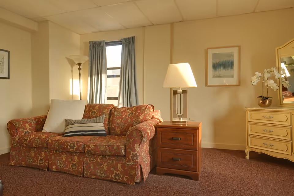 A cozy living room area with a floral patterned sofa adorned with two pillows, one white and one striped. Next to the sofa is a wooden side table with a modern lamp on it. Behind the sofa is a window with blue curtains, and a tall floor lamp stands in the corner. On the right side, there is a cream-colored dresser with a mirror and a white orchid plant on top. A framed abstract painting hangs on the wall above the dresser.