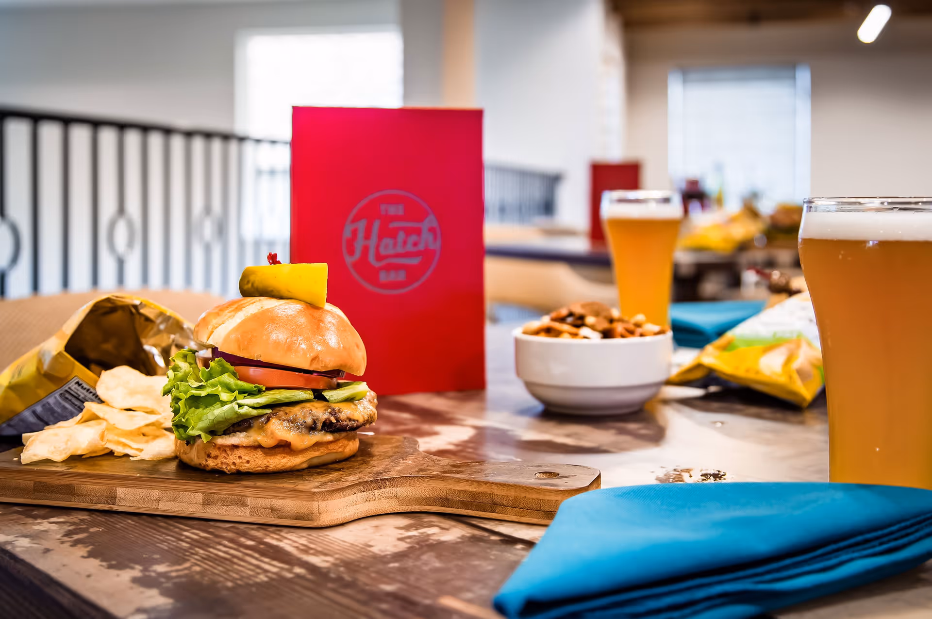 A cheeseburger with chips on a wooden board beside beers, a bowl of nuts and a red menu on a dining table.