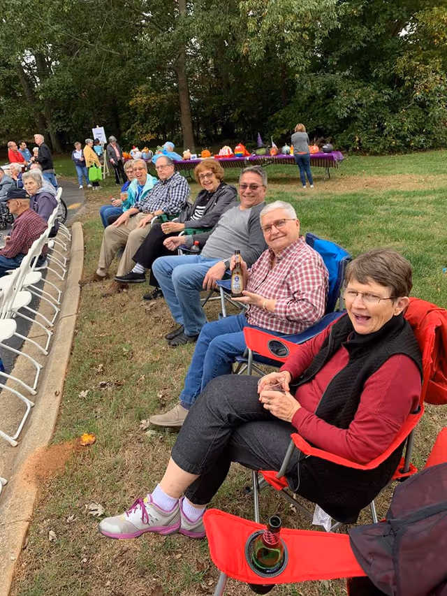 A group of elderly people sitting outdoors in folding chairs on a grassy area, smiling and enjoying drinks. Behind them, there is a table covered with a purple cloth displaying various decorated pumpkins. Trees and more people are visible in the background.