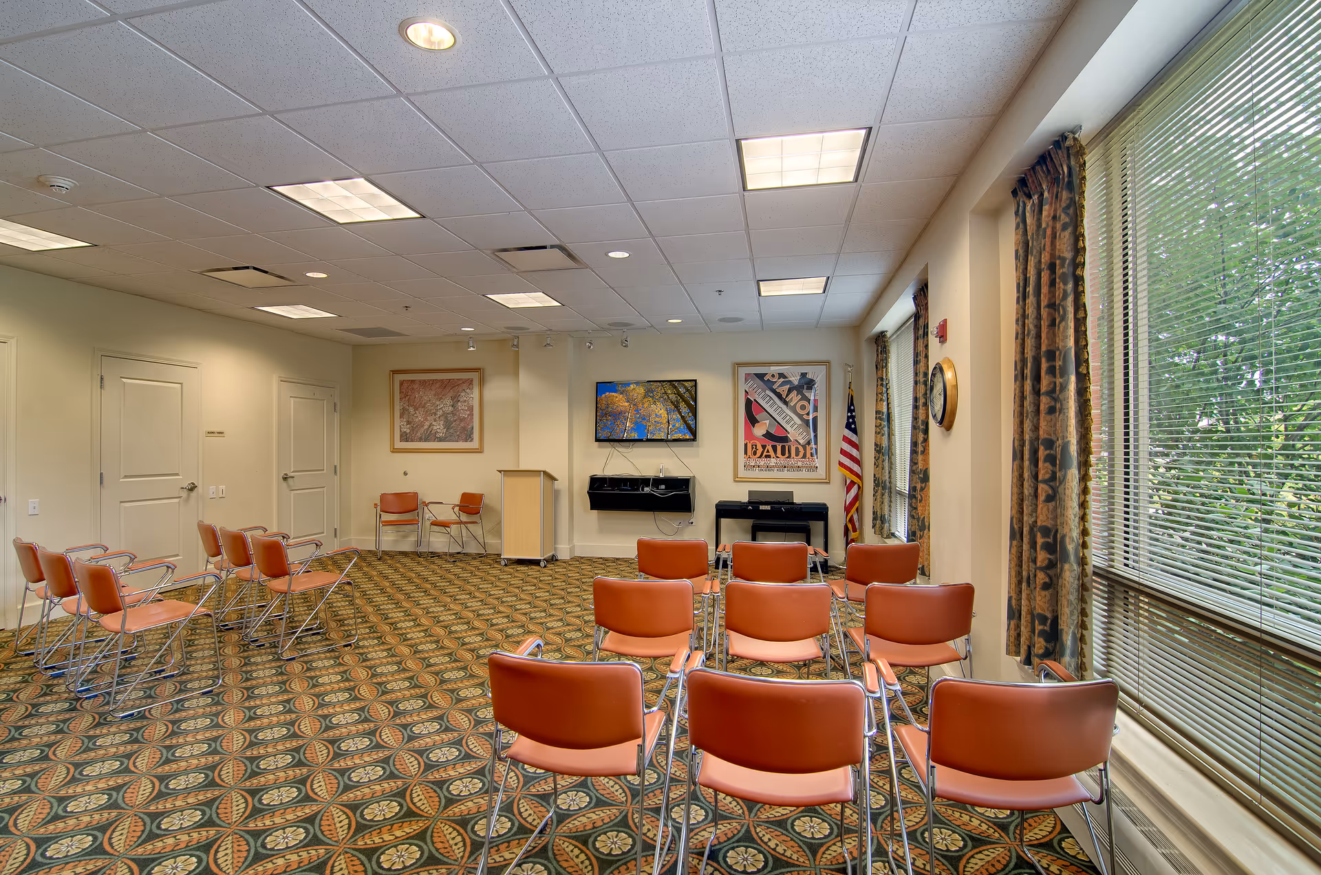 A medium-sized meeting or activity room with rows of orange chairs facing a wall with framed artwork, a podium, and an electronic piano. The room has patterned carpet, large windows with floral curtains, and an American flag in the corner.