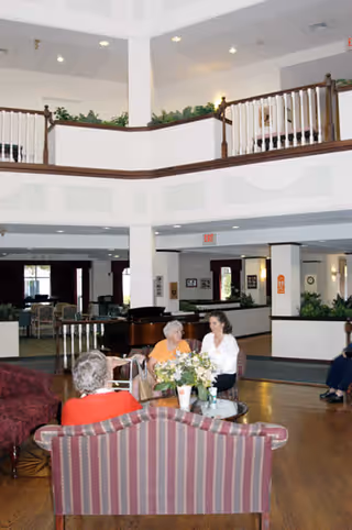 A spacious senior living facility common area with high ceilings and a balcony railing above. Several elderly people are seated on sofas and chairs around a coffee table with a flower arrangement. The room has wooden flooring and is decorated with plants and framed pictures on the walls.