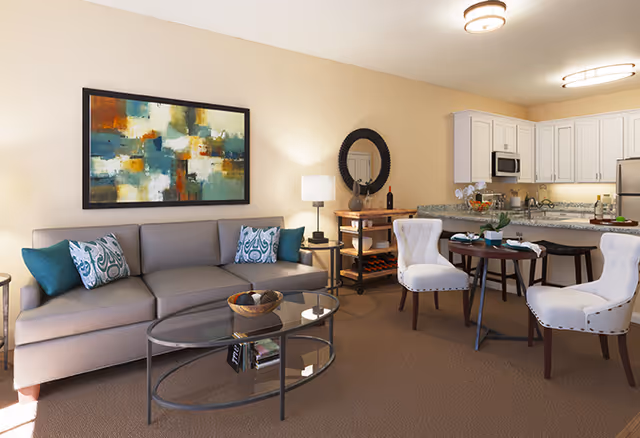 A cozy living area in a senior living facility featuring a gray sofa with decorative pillows, a glass coffee table, a round dining table with four white upholstered chairs, and a kitchen area with white cabinets, granite countertops, and stainless steel appliances. The room is warmly lit with a table lamp and ceiling lights, and decorated with a colorful abstract painting and a round mirror above a wooden console table.