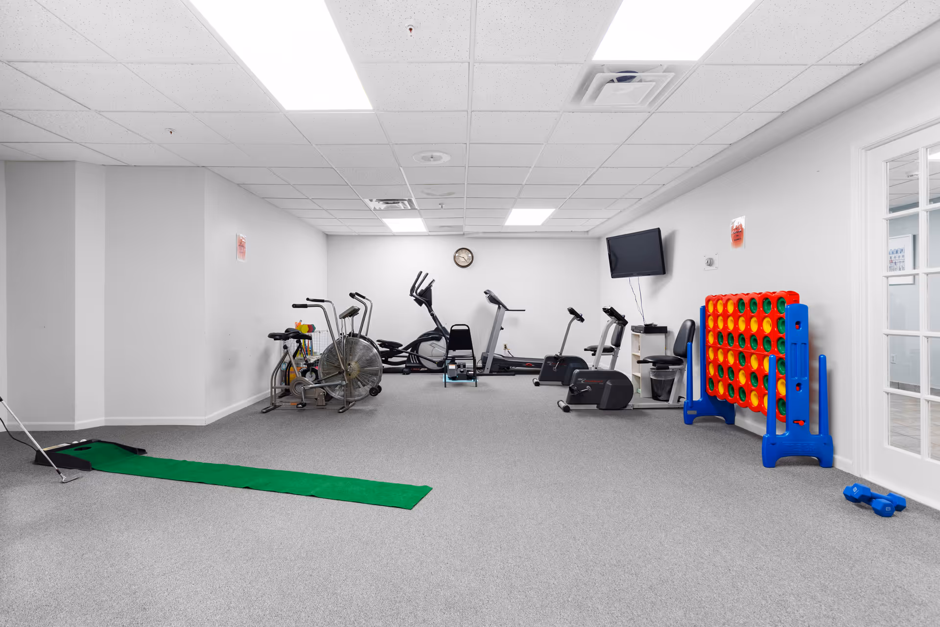 A fitness and recreation room with exercise equipment including stationary bikes, elliptical machines, and a treadmill. There is a green putting mat for golf on the floor, a large Connect Four game against the wall, a wall-mounted TV, and a pair of blue dumbbells on the floor near a glass door.