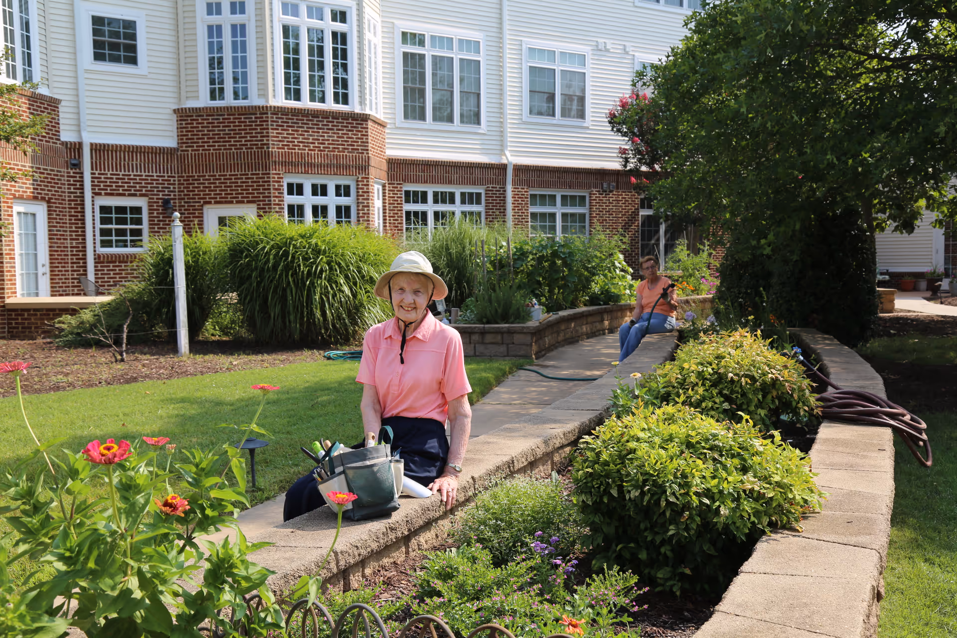An elderly woman wearing a pink shirt and a sun hat is sitting on a low stone wall in a garden area outside a brick and white siding building. She has gardening tools and a bag beside her. Another person is sitting further back on the same wall, holding a garden hose. The garden is lush with green bushes and colorful flowers.