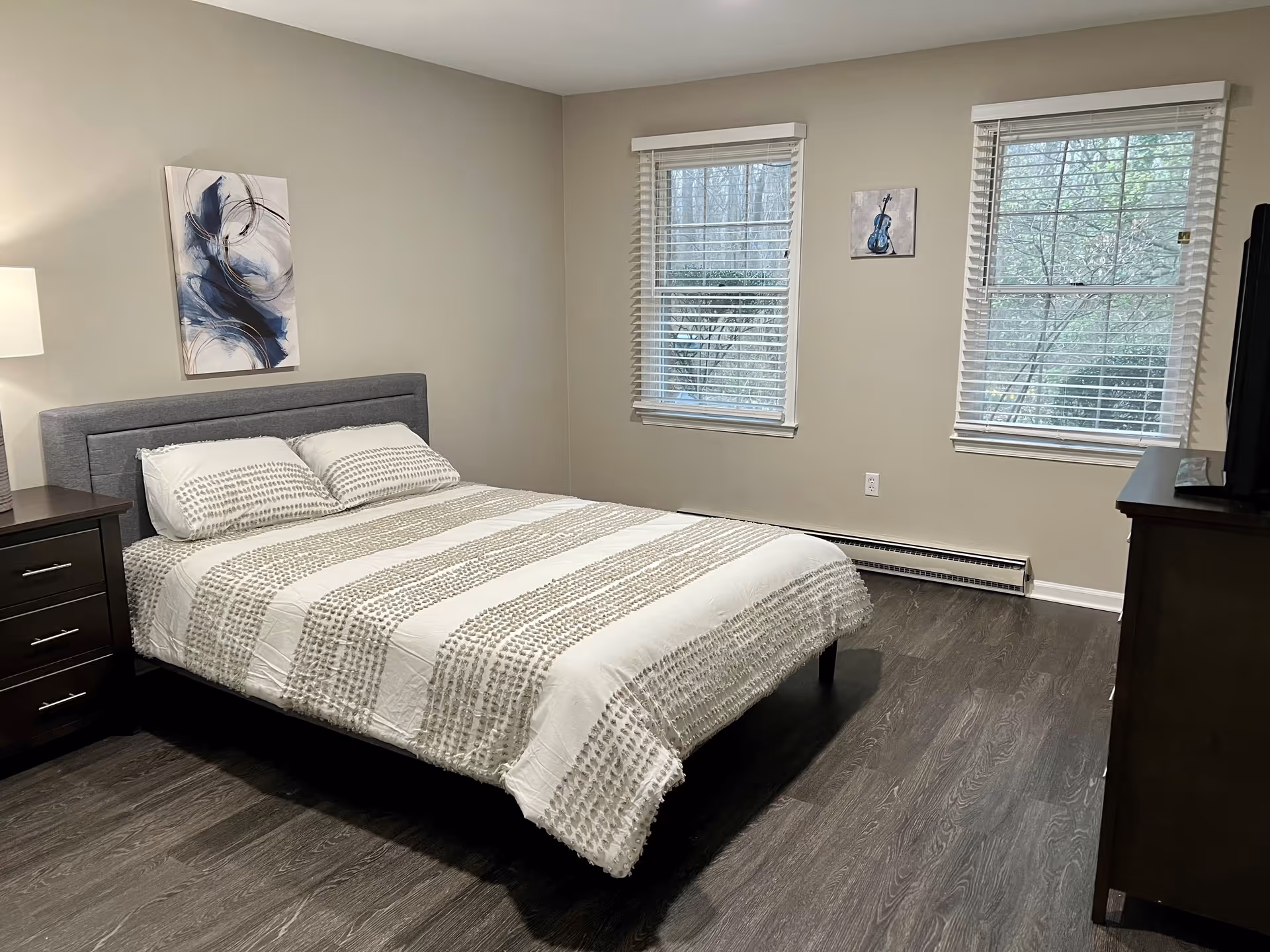 A bedroom with a neatly made bed featuring a white and gray striped comforter and two pillows. There is a dark wooden nightstand with a lamp on the left side of the bed. Two windows with white blinds are on the wall opposite the bed, and a dark wooden dresser with a TV on top is on the right side. The walls are painted beige and have two pieces of artwork hanging. The floor is a dark wood laminate.