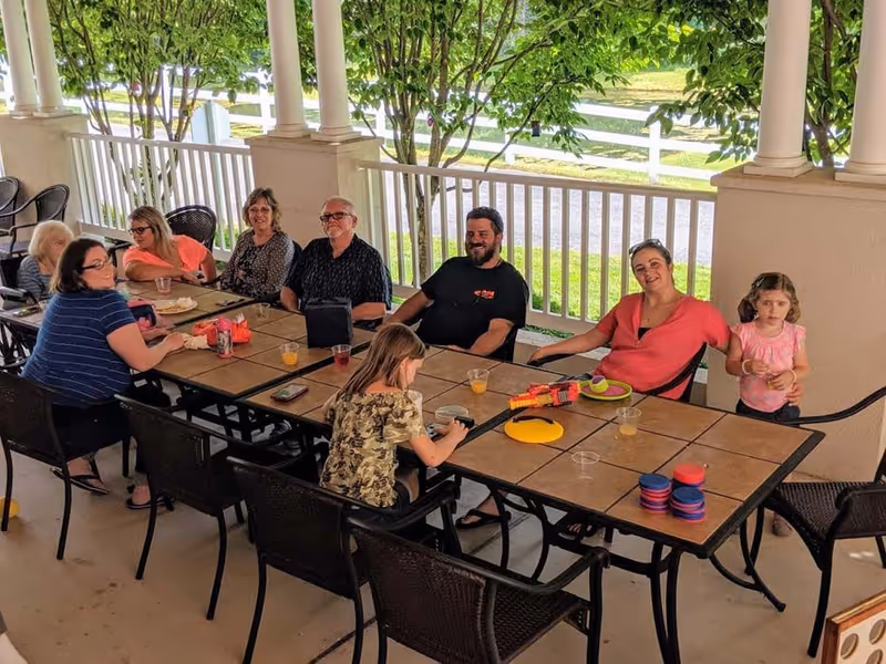 A group of people, including adults and children, sitting and standing around a long outdoor table on a covered porch with white railings and columns. The table has drinks, plates, and toys on it. Green trees and a white fence are visible in the background.