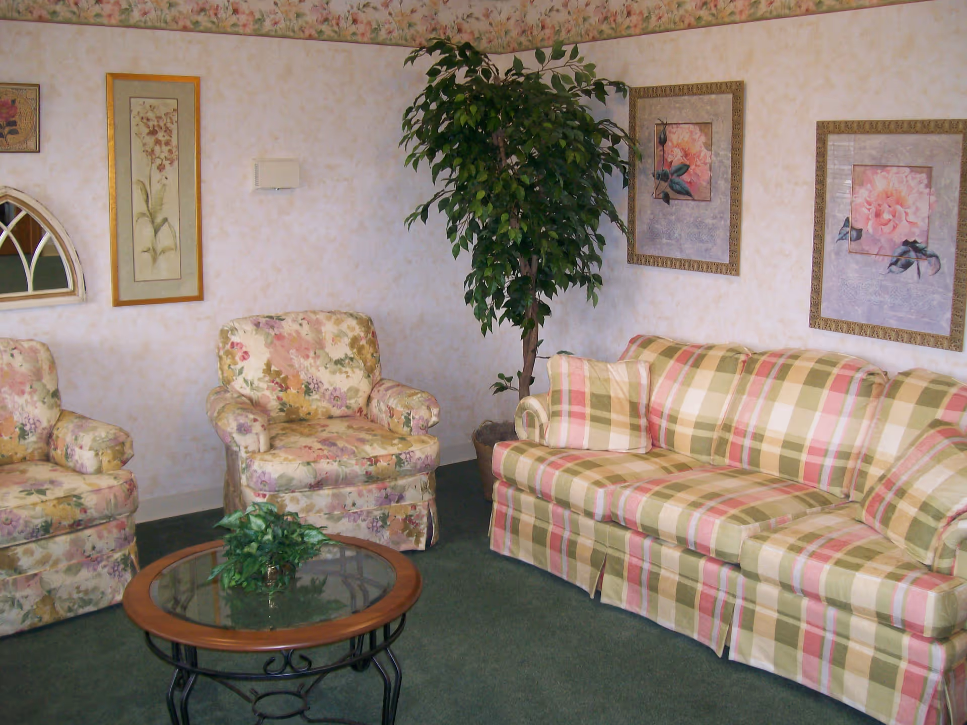 A cozy living room with floral and plaid upholstered furniture including a sofa and two armchairs. There is a round glass-top coffee table with a small green plant on it. The walls are decorated with floral paintings and a large potted plant is placed in the corner.