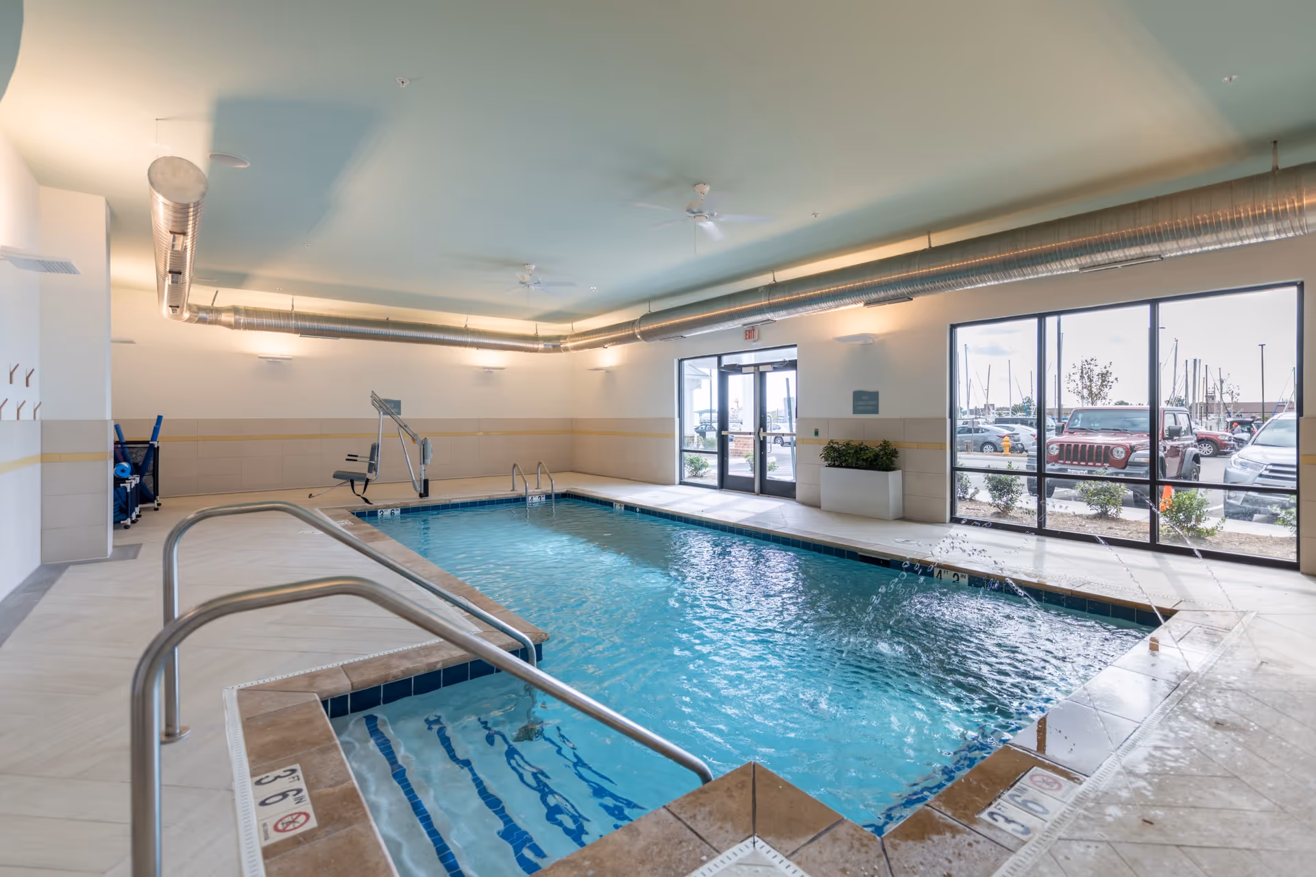 Indoor swimming pool area with clear blue water, a small attached hot tub, metal handrails, and a pool lift chair. Large windows and glass doors provide natural light and a view of parked cars outside. The ceiling has exposed ductwork and ceiling fans.