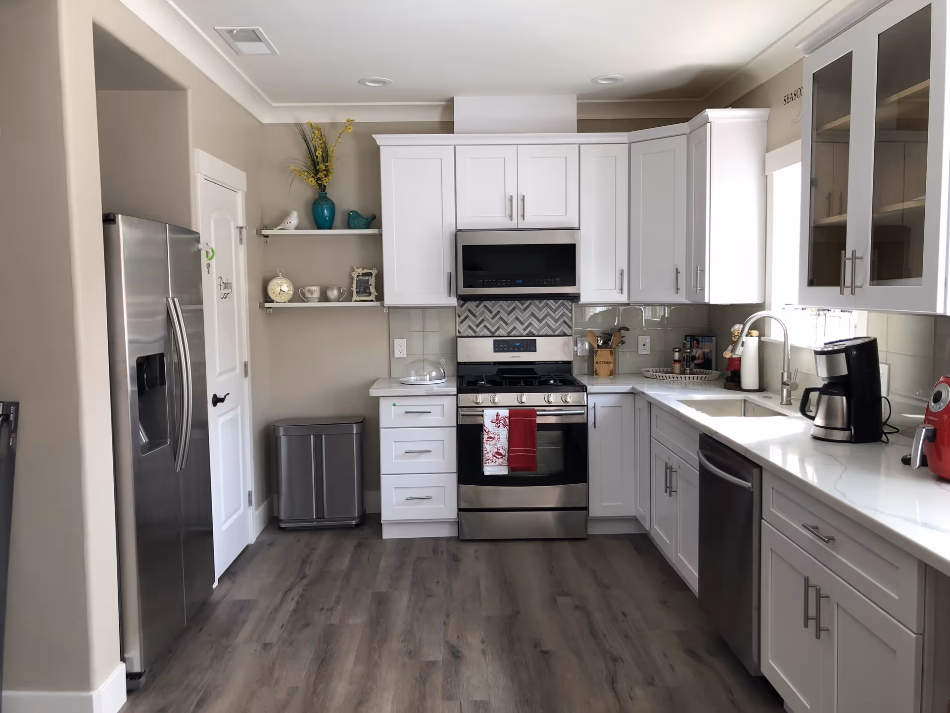 Modern kitchen with white cabinets, stainless steel appliances including a refrigerator, stove, microwave, and dishwasher. The kitchen has a light-colored countertop, a coffee maker, and a red air fryer on the counter. There are decorative items on shelves and a trash bin near a pantry door.