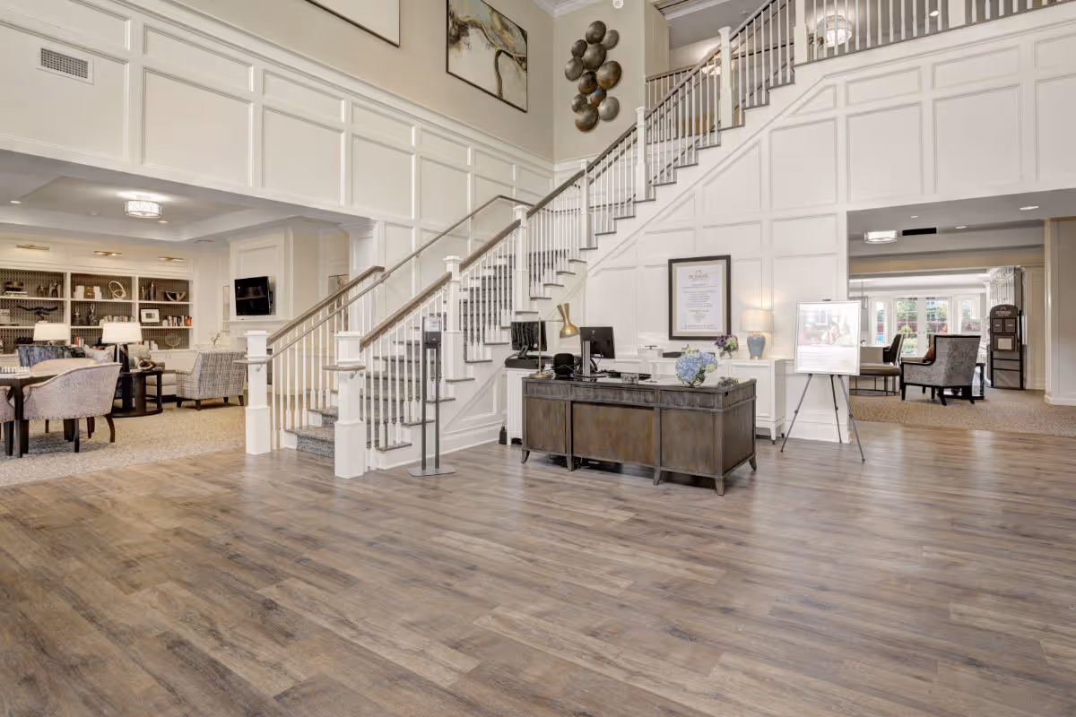 Spacious and well-lit senior living facility lobby with wooden flooring, a reception desk, a staircase with white railings, and comfortable seating areas with armchairs and tables. The walls are decorated with artwork and paneling, and there is a bookshelf with various items in the background.