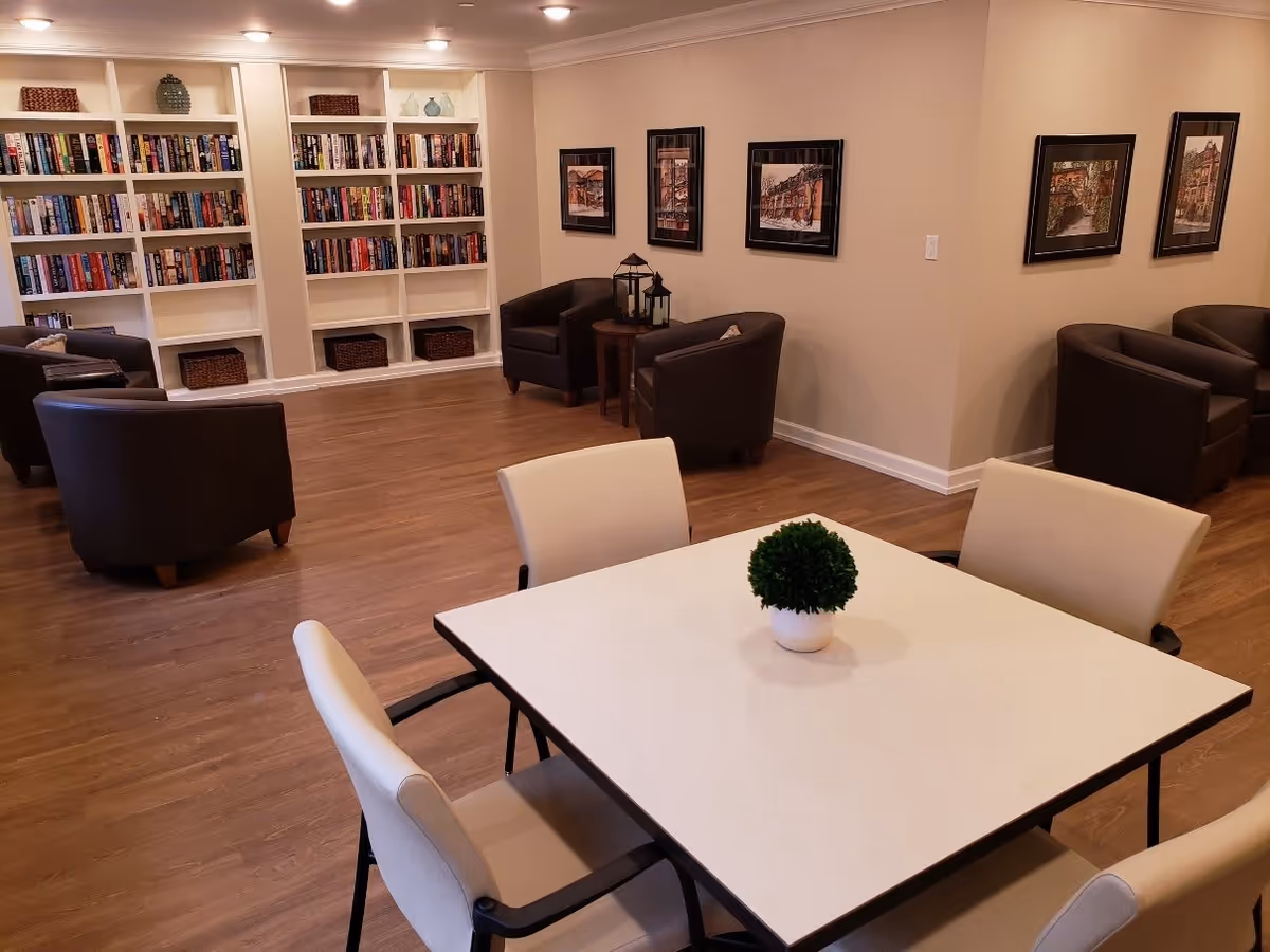 A cozy common area with a white square table surrounded by four beige chairs, a small green potted plant on the table, several dark brown armchairs arranged around the room, built-in white bookshelves filled with books and decorative items, and framed artwork on beige walls.