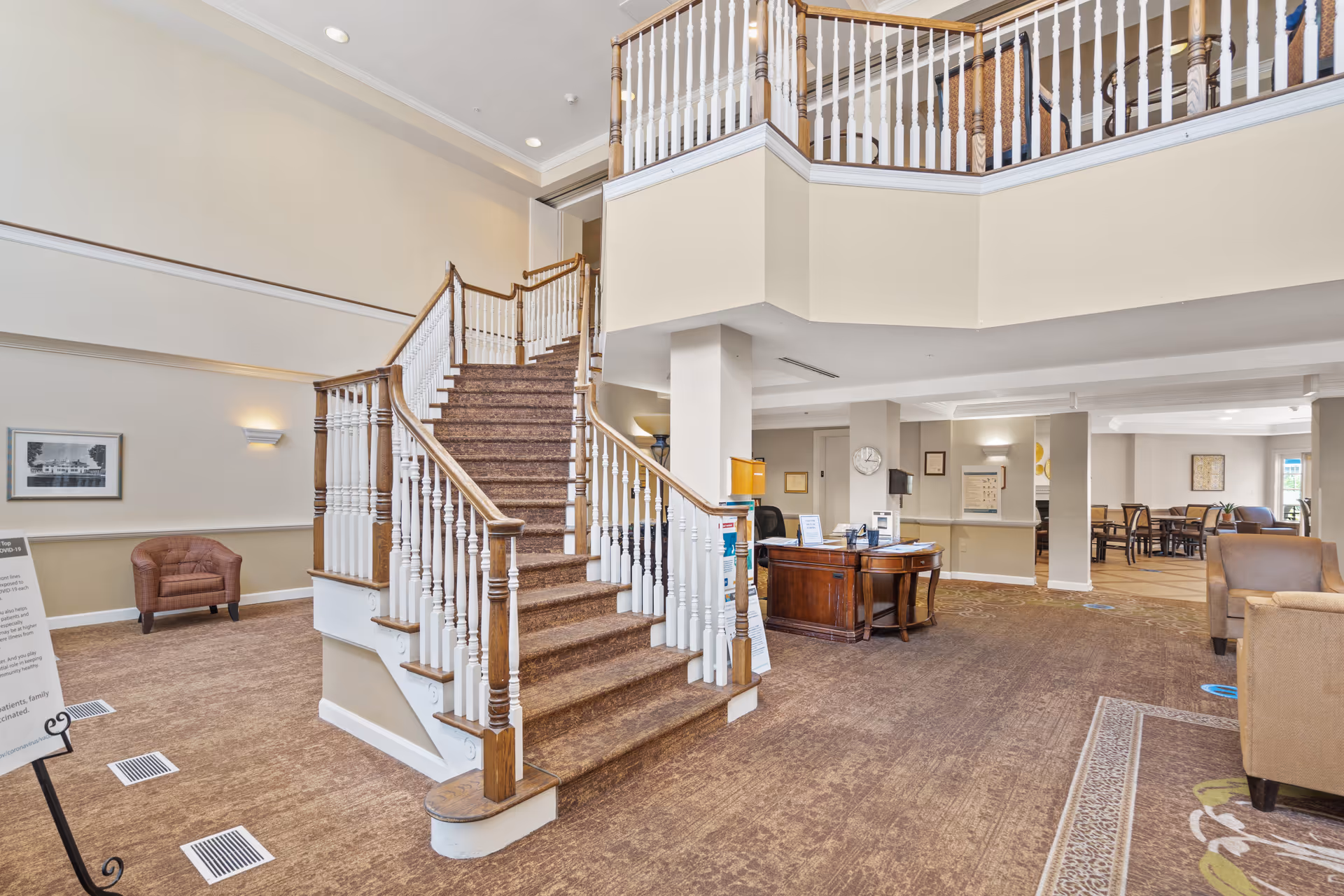 Interior view of a senior living facility lobby featuring a central staircase with wooden handrails and white balusters. The space includes a reception desk, seating areas with armchairs, and a dining area visible in the background. The decor is warm with beige walls and carpeted floors.