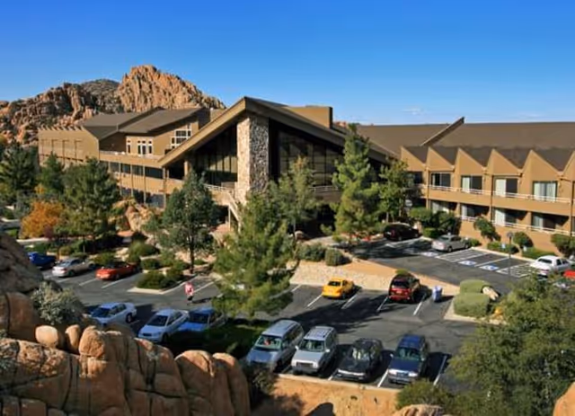Exterior view of Granite Gate Senior Living facility showing a large two-story building with a stone facade and multiple windows, surrounded by trees and rocky landscape. Several cars are parked in the parking lot in front of the building under a clear blue sky.