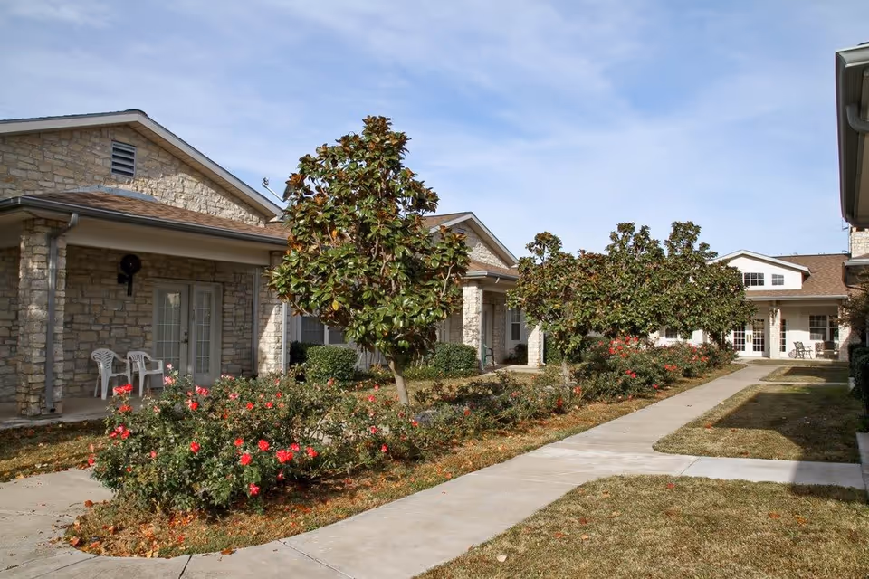 Outdoor courtyard area of a senior living facility with stone buildings, small trees, and flowering bushes along a concrete walkway. White chairs are placed on the porches of the buildings under a clear sky.