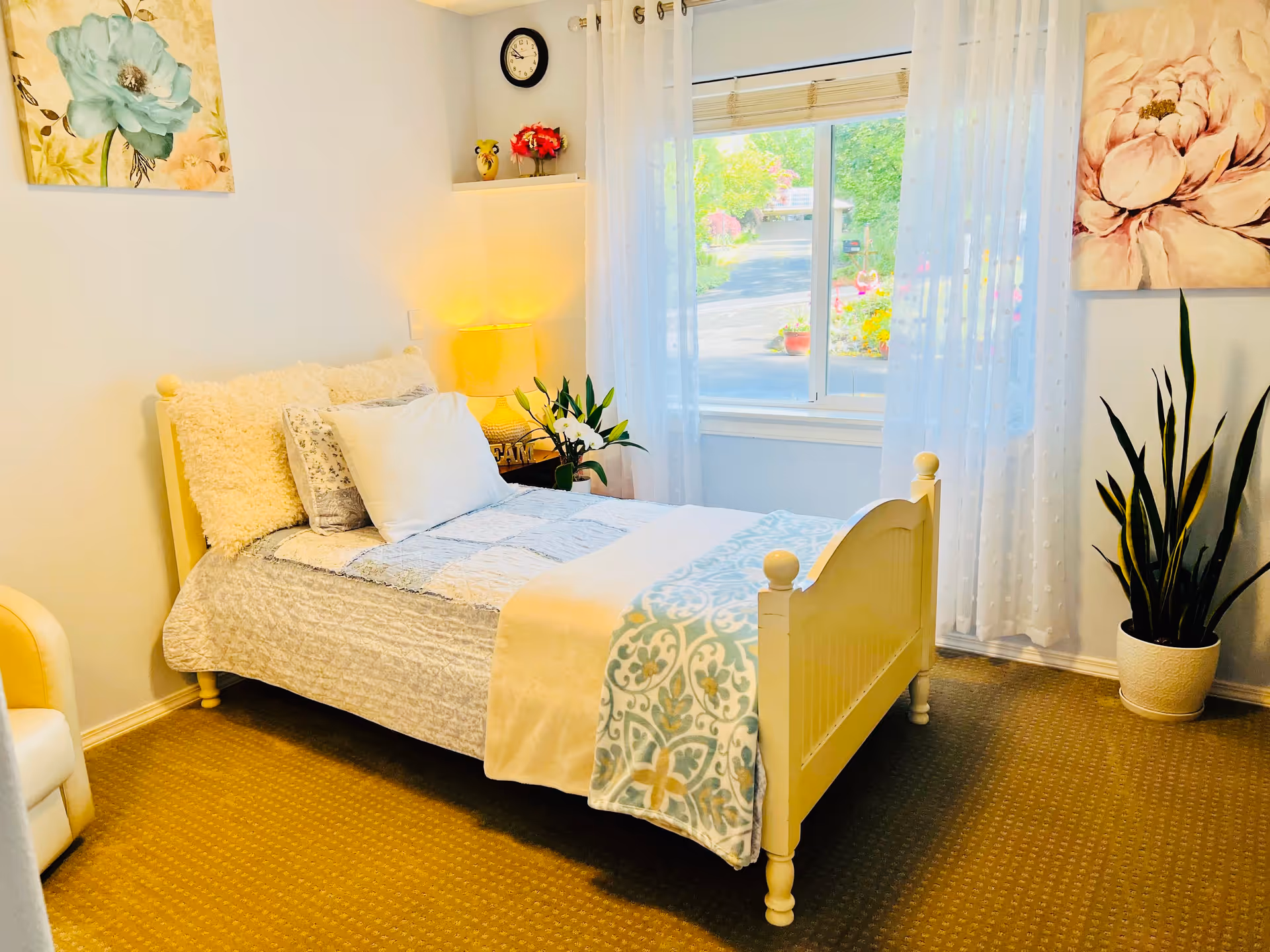 A cozy bedroom with a single cream-colored bed adorned with multiple pillows and a light blue patterned blanket. A nightstand with a lamp and a small plant sits next to the bed. The room has a window with sheer white curtains letting in natural light, and there are two floral paintings on the walls. A potted plant is placed on the floor near the window.