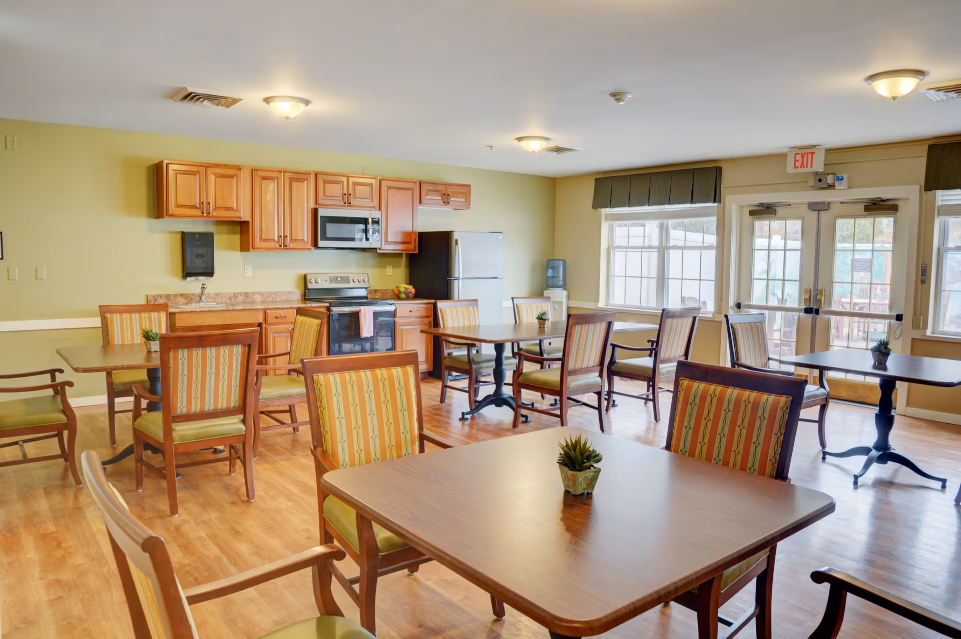 A bright dining room with several wooden tables and chairs featuring striped upholstery. The room has a kitchen area with wooden cabinets, a microwave, stove, and refrigerator. Large windows and glass doors allow natural light to fill the space, and small potted plants decorate the tables.