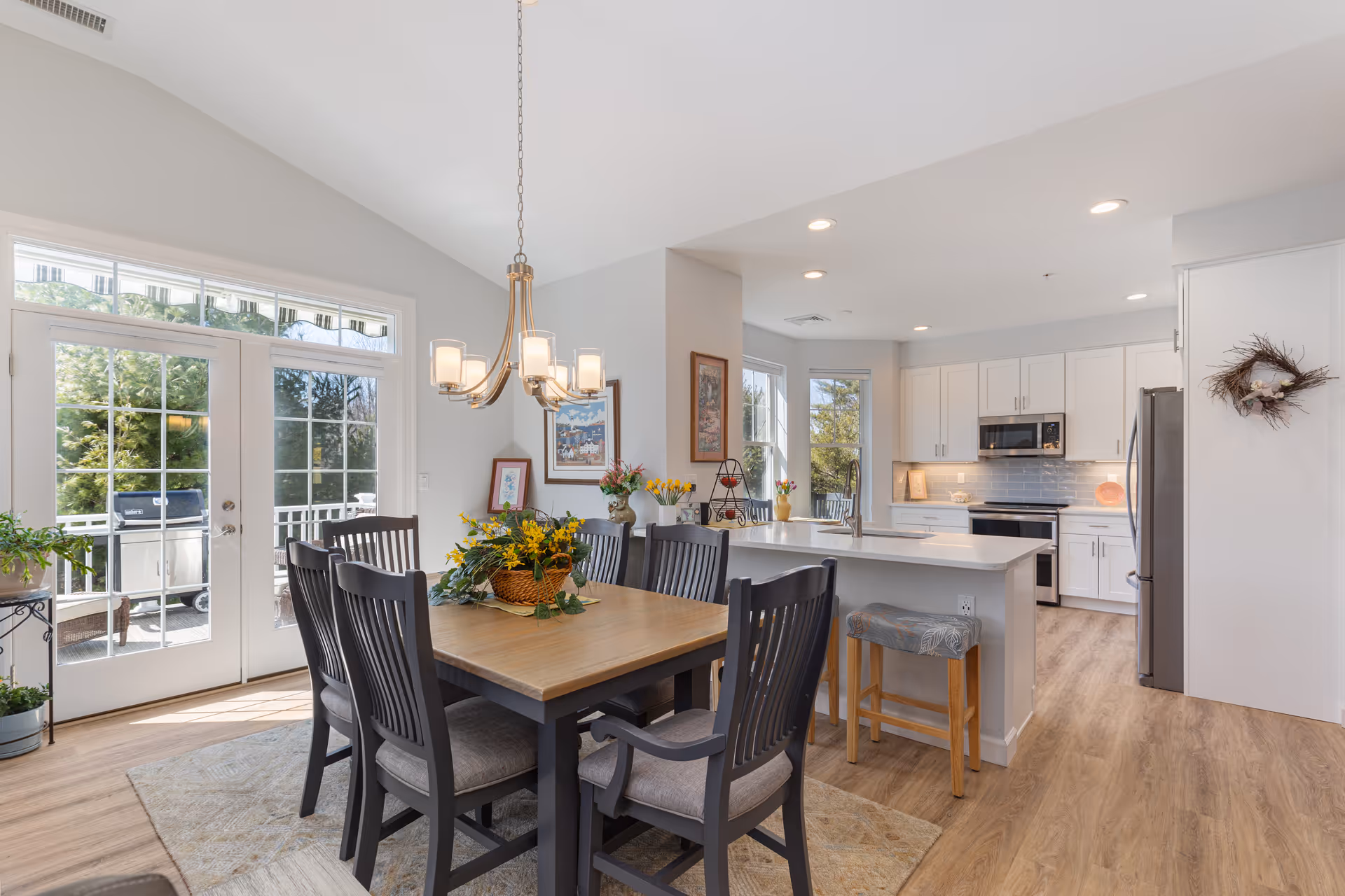 Bright open dining area with a wooden table and chairs adjacent to a modern white kitchen and island, with glass doors leading to an outdoor patio.
