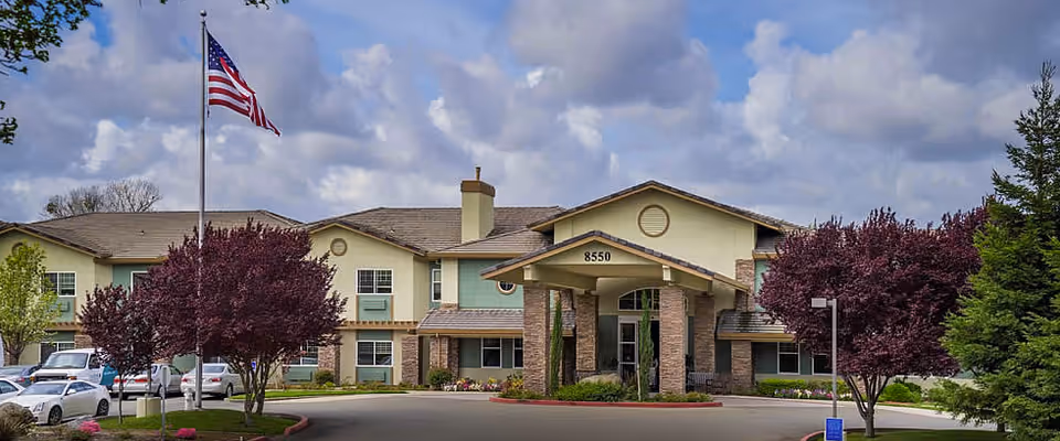Front exterior view of Eskaton Granite Bay facility with a covered entrance, several windows, an American flag on a flagpole, and landscaped trees and shrubs under a partly cloudy sky.