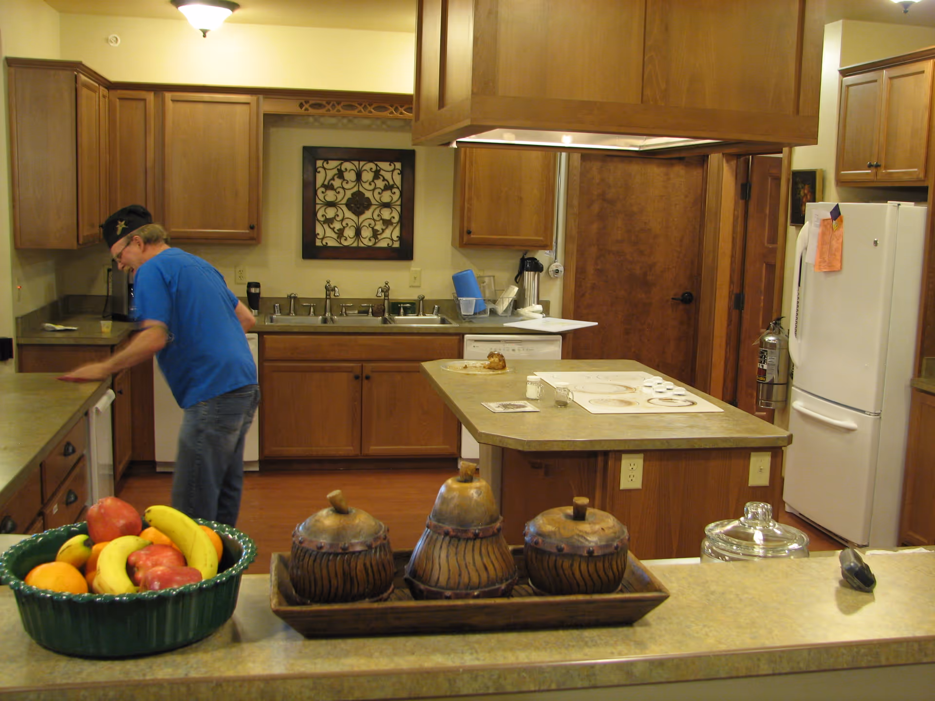 A kitchen with wooden cabinets, a white refrigerator, a stove on an island counter, and a man in a blue shirt cleaning the countertop. There is a bowl of fruit and a set of decorative wooden containers on the counter in the foreground.
