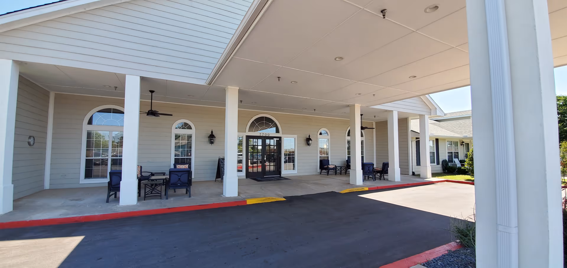Entrance of Hefner Mansions Senior Independent Living facility with a covered drop-off area supported by white columns. There are several seating areas with chairs and tables on the porch, large windows with arched tops, and a double glass door entrance. The driveway is paved and bordered by red and yellow painted curbs.