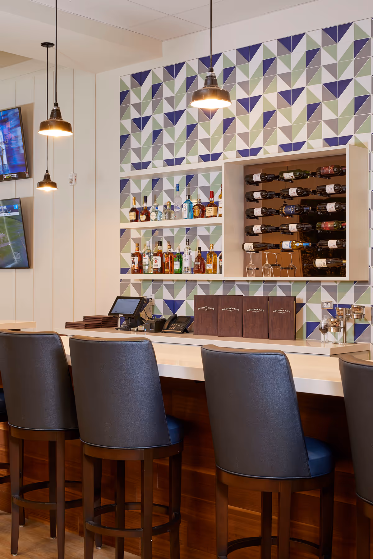 A modern bar area with four bar stools, pendant lights, a geometric tiled wall, shelves of liquor bottles and a wine rack.