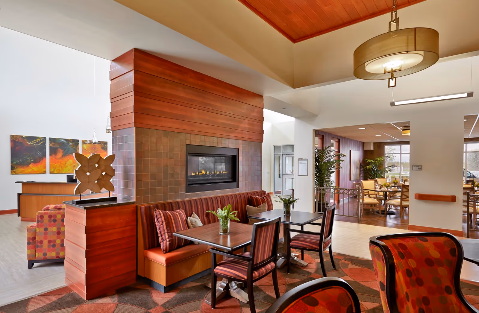 A cozy seating area in a senior living facility with a modern fireplace set in a tiled and wooden wall. The space features a built-in bench with striped cushions, two wooden tables with matching striped chairs, and small green plants on the tables. In the background, there is a dining area with round tables and chairs, large windows letting in natural light, and decorative artwork on the walls. The ceiling has a wooden finish with a hanging light fixture.